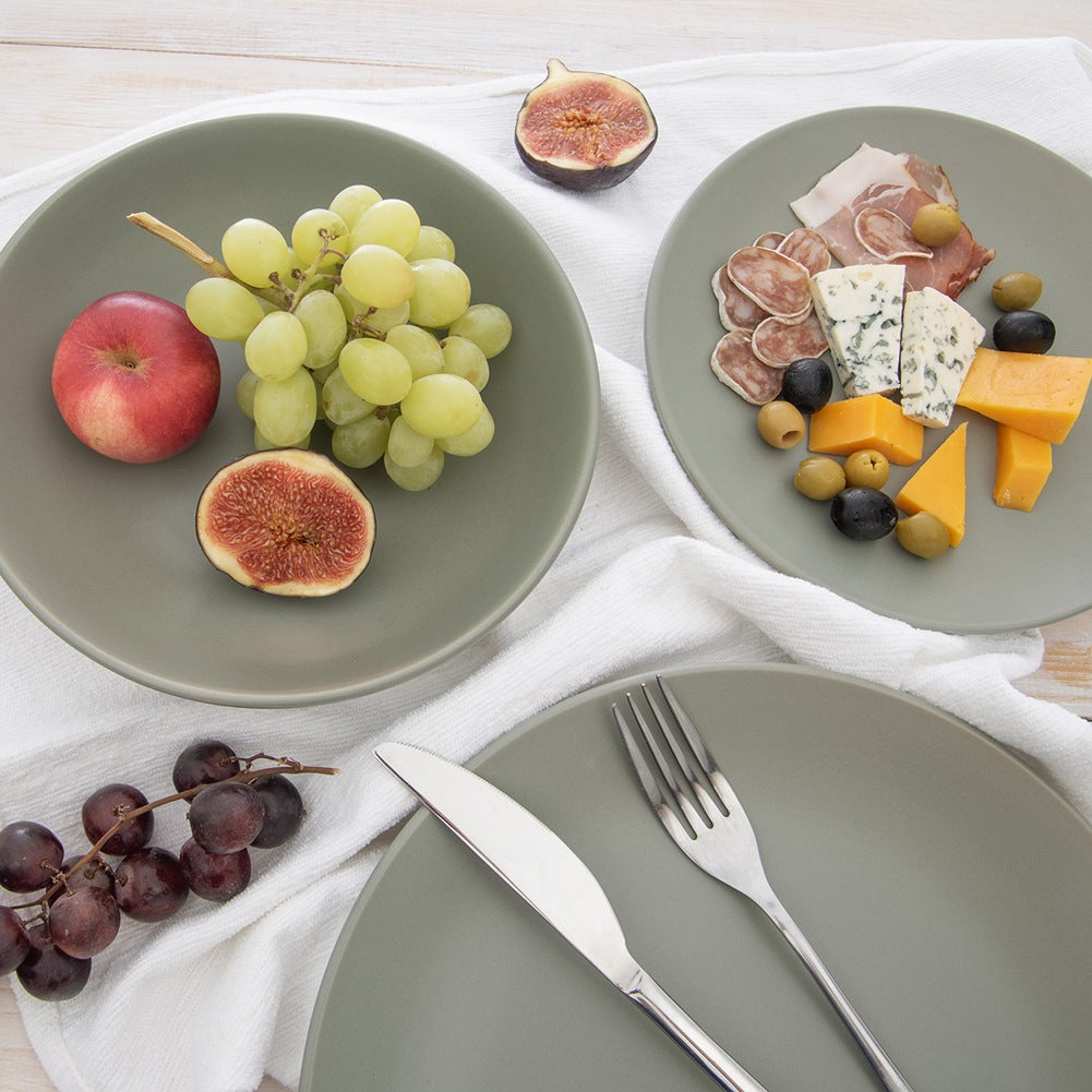 Platos con comida: uvas, manzana, higos, queso, aceitunas y embutidos. Cubiertos en un plato. Paño blanco sobre madera.