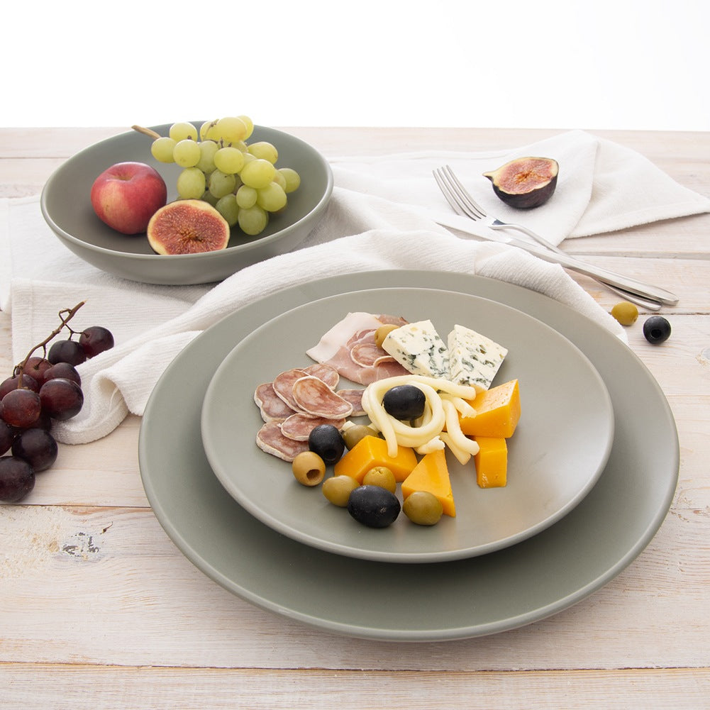 Un plato con queso, aceitunas y carne está en una mesa de madera. Un cuenco tiene uvas, un melocotón y un higo.