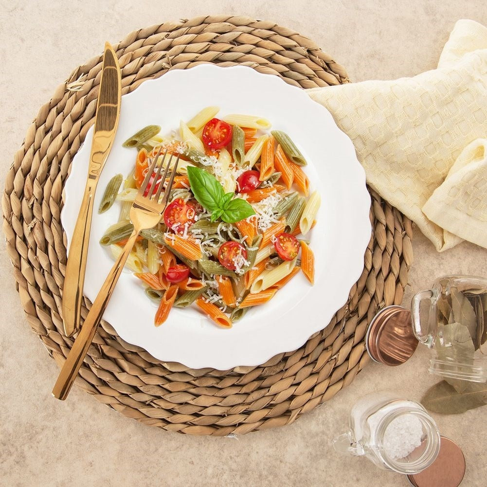 Plato de pasta con tomates y albahaca en un plato blanco, cubertería dorada, sobre una esterilla tejida. Ingredientes y tela al fondo.