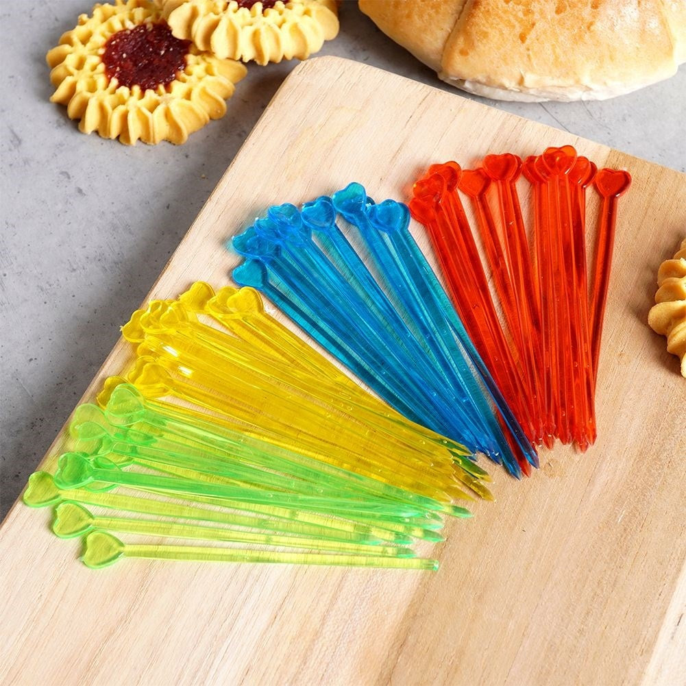 Una tabla de madera con brochetas coloridas con forma de corazón en verde, amarillo, azul y rojo. Galletas y pan al fondo.