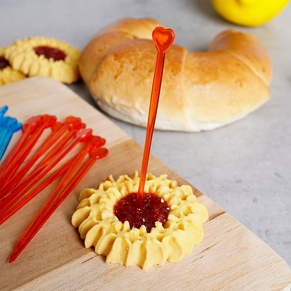 Una galleta con relleno rojo es atravesada por un pincho con forma de corazón. Otras galletas, un cruasán y pinchos están en el fondo.