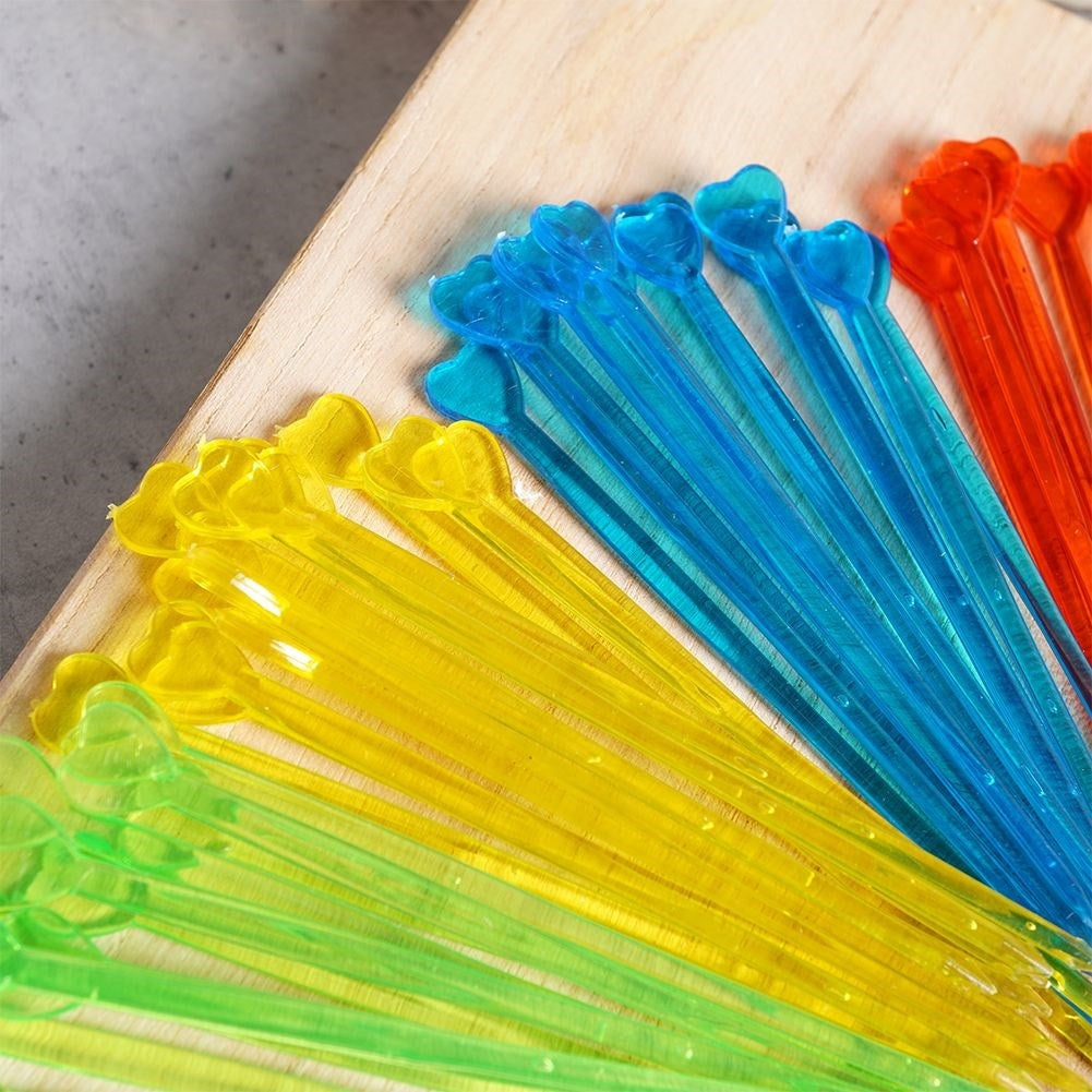 Brochetas coloridas con forma de corazón sobre una tabla de madera. Las brochetas son amarillas, verdes, azules y naranjas. El fondo es gris.