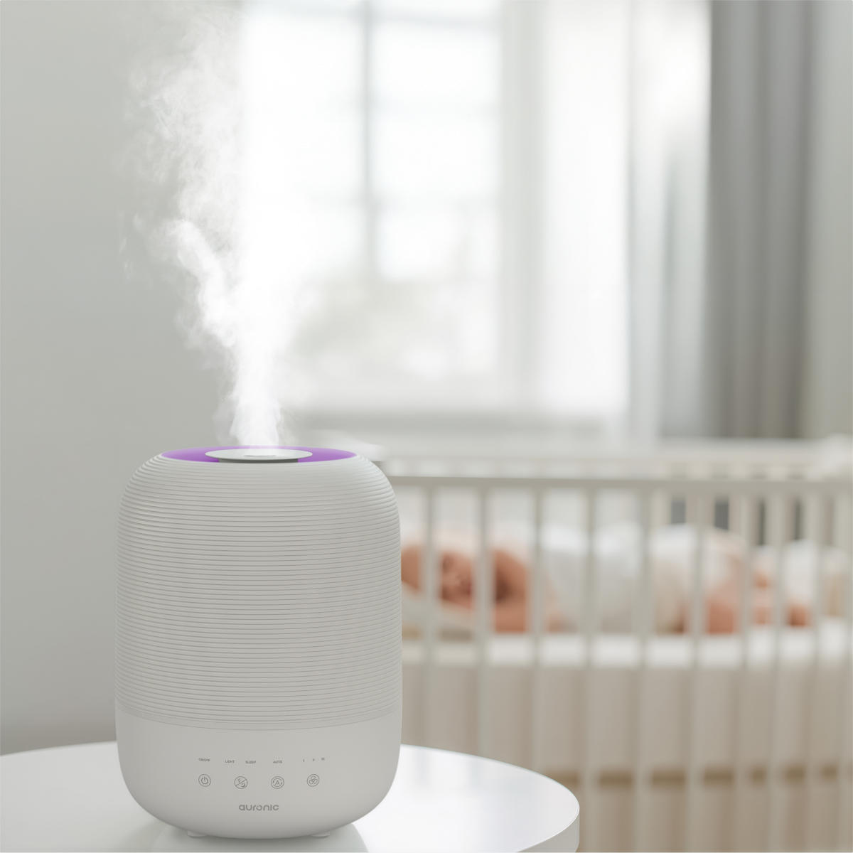 A white humidifier with purple top emits steam. A baby sleeps in a crib in the background. The scene is bright and airy.