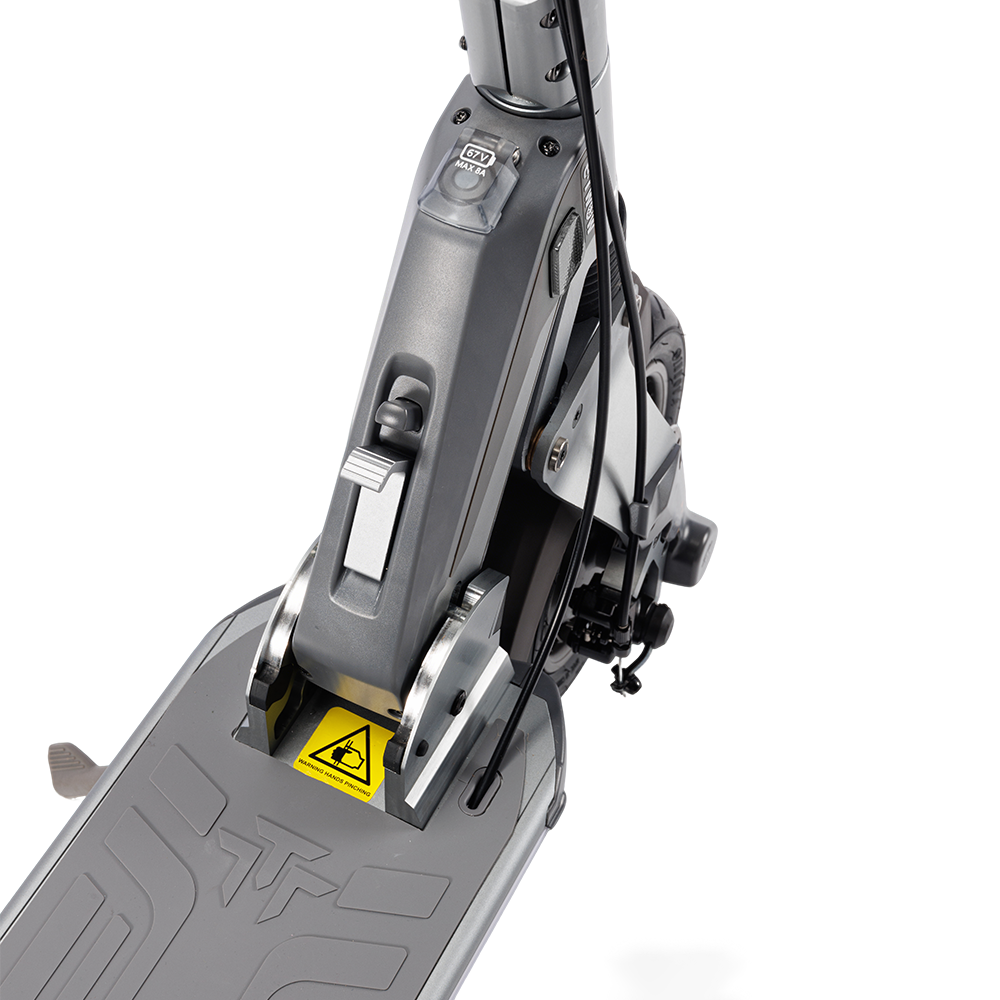 Zbliżenie szarej hulajnogi elektrycznej. Widoczny mechanizm składania, etykieta ostrzegawcza oraz część przedniego koła i kierownicy.