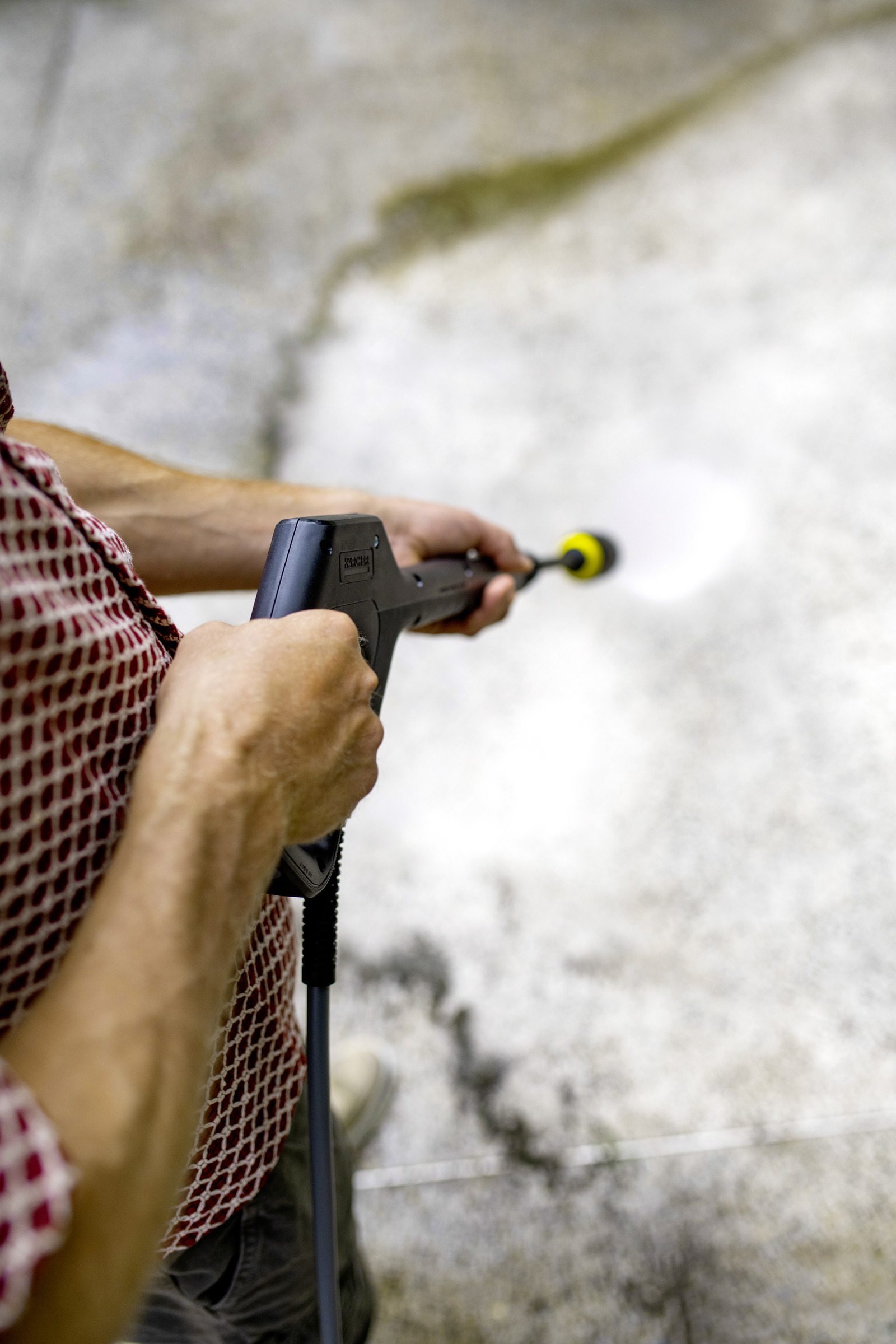 Osoba trzyma myjkę ciśnieniową, spryskując wodą betonową powierzchnię. Ma na sobie czerwoną koszulę we wzory.