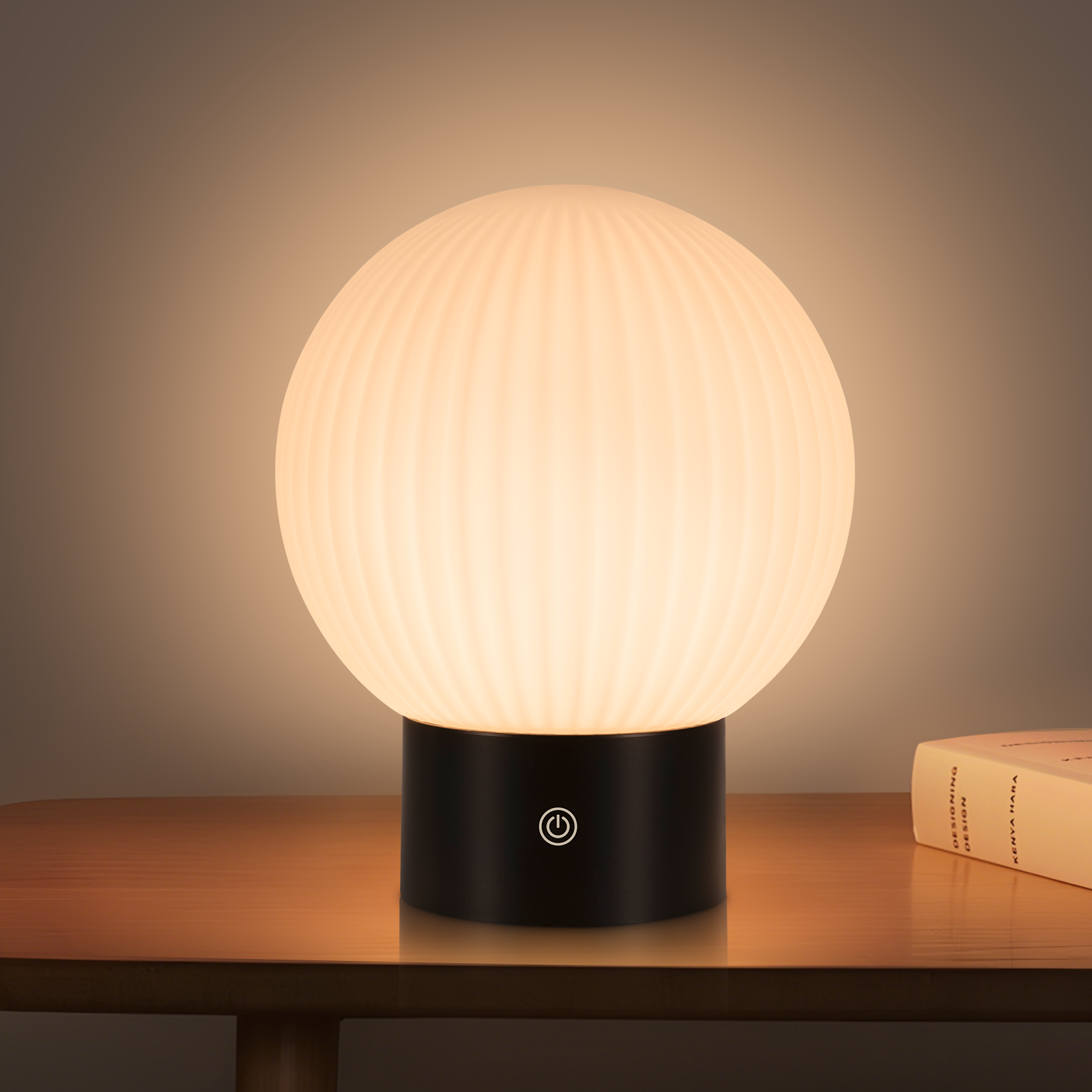Świecąca lampa w kształcie kuli z czarną podstawą stoi na drewnianej powierzchni. Książka jest częściowo widoczna po prawej stronie.