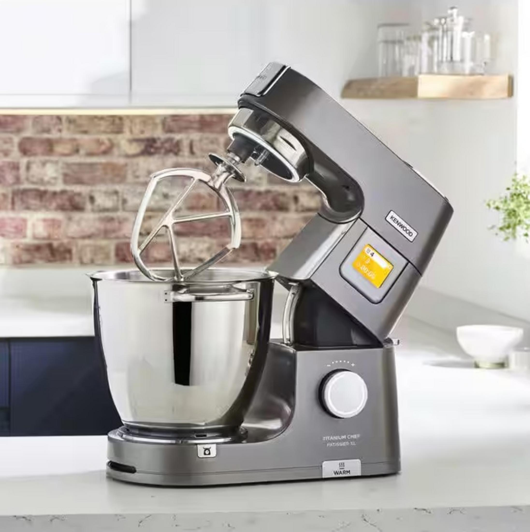 Ein grauer Kenwood-Küchenmixer mit einer Edelstahlschüssel und einem Schläger steht auf einer weißen Theke. Im Hintergrund eine Backsteinmauer.