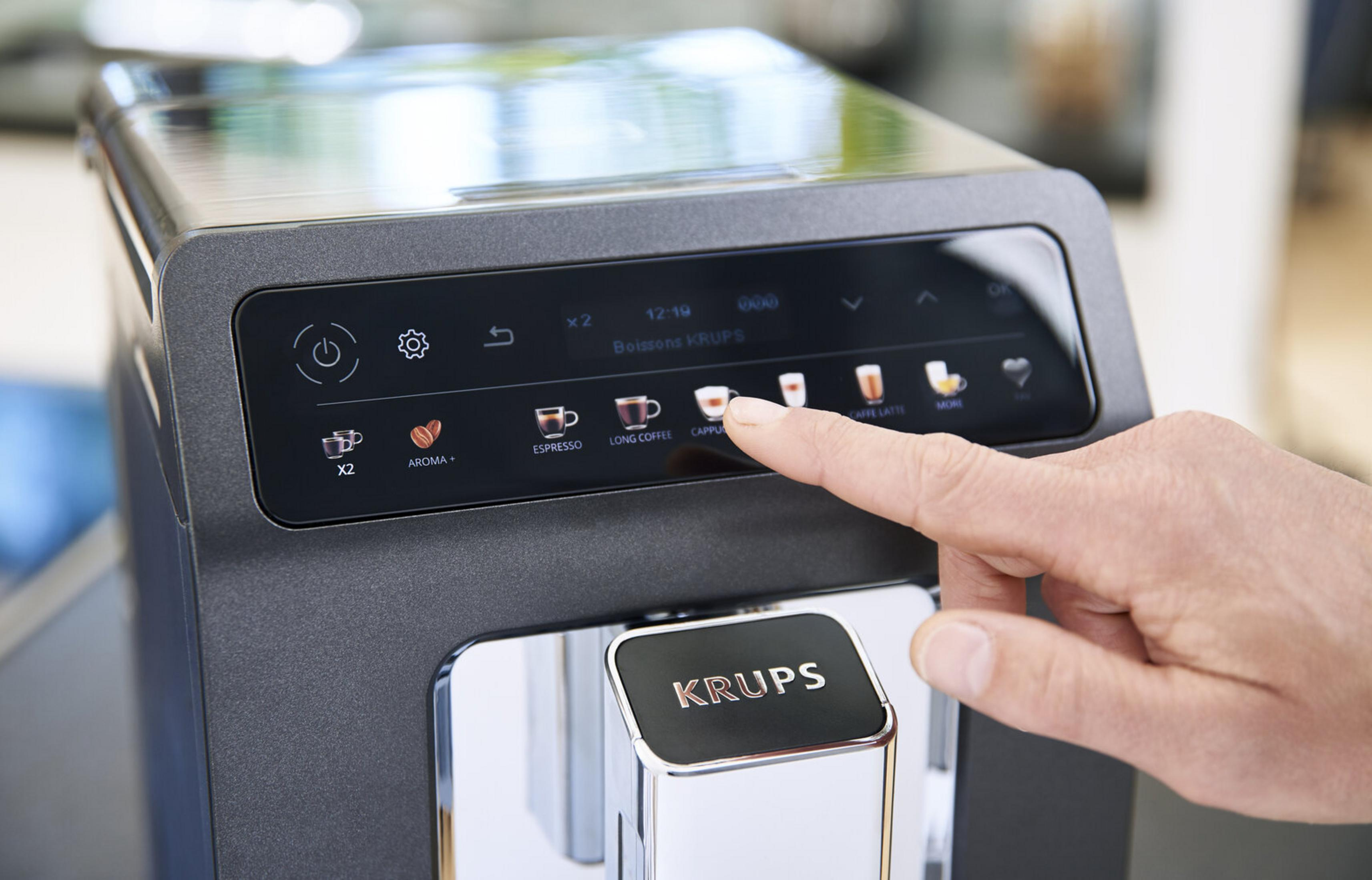 Ręka naciska opcję cappuccino na cyfrowym wyświetlaczu ekspresu do kawy Krups. Maszyna jest czarna i srebrna.