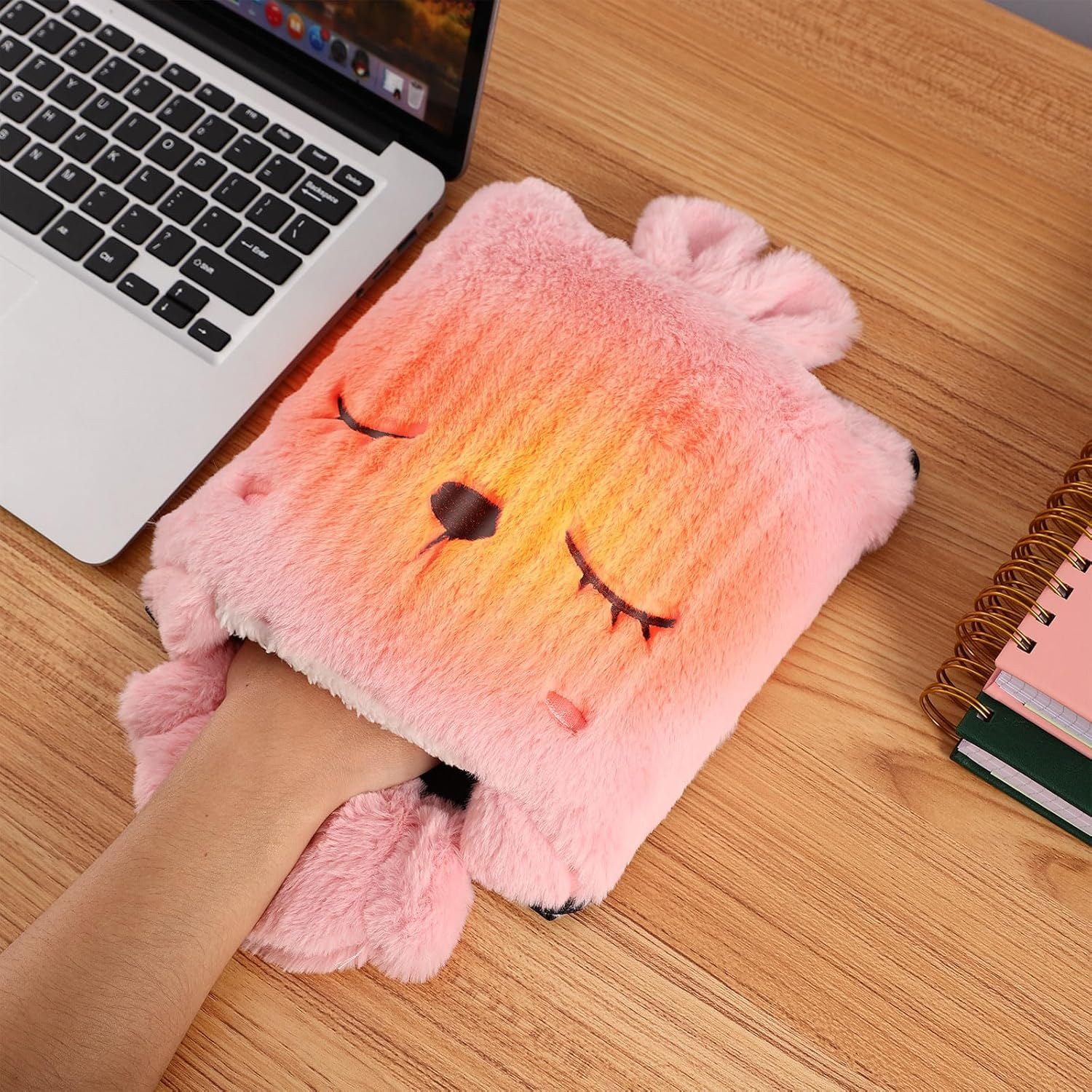 Eine Hand in einem rosa Plüsch-Handwärmer in Form eines Gesichts auf einem Holztisch. Ein Laptop und Notizbücher sind in der Nähe.