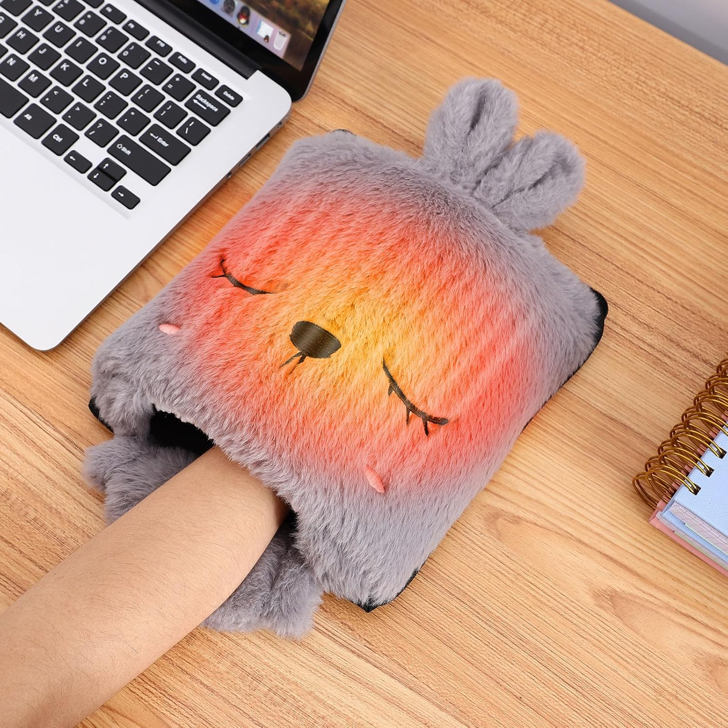 Eine Hand in einem grauen, flauschigen Handwärmer in Fuchsform, in der Nähe eines Laptops und eines Notizblocks auf einem Holztisch.