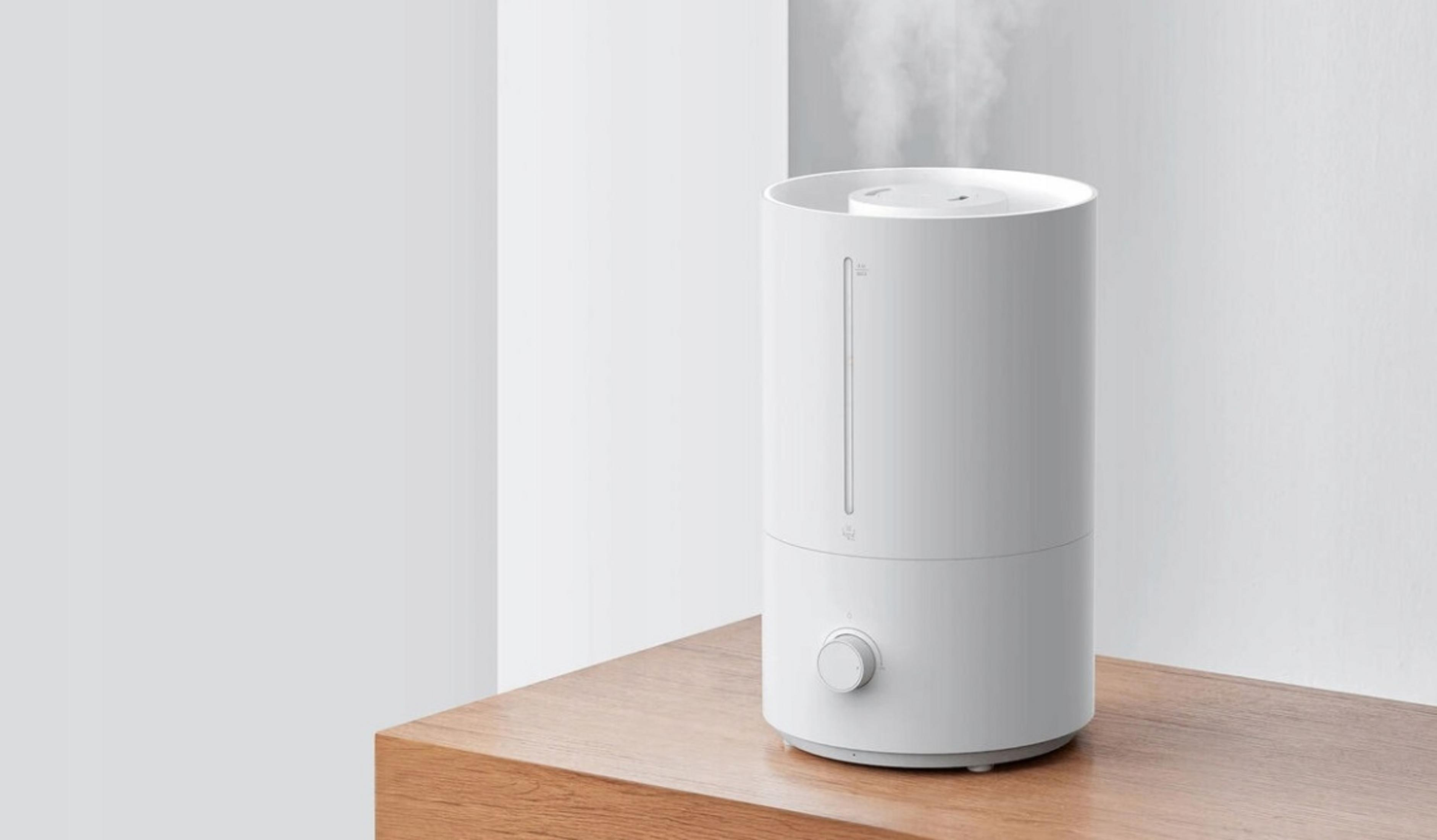 Biały nawilżacz na drewnianym stole. Ma pokrętło i wskaźnik poziomu wody. Para unosi się z góry na białym tle.