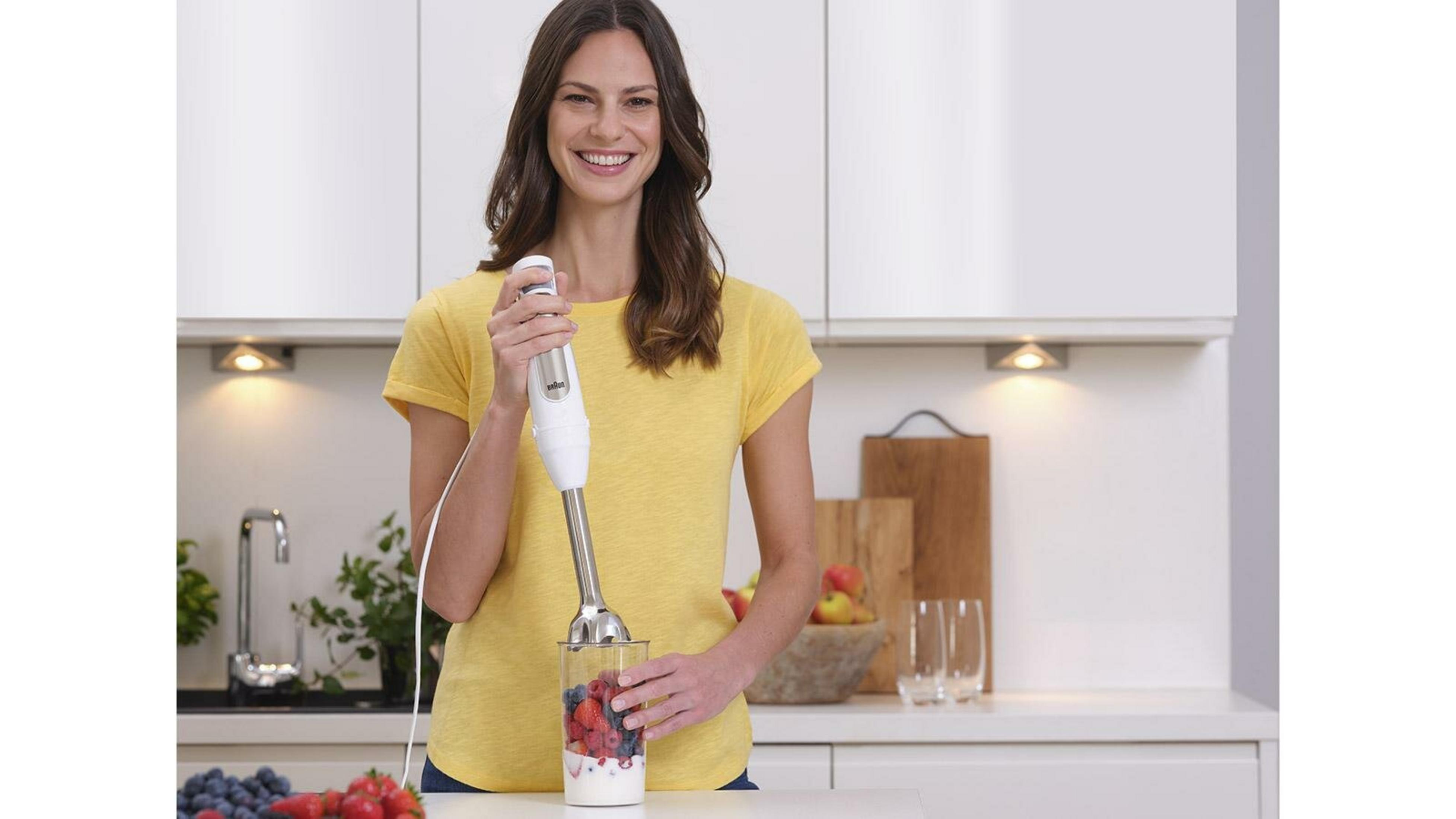 Una mujer con camisa amarilla sonríe, sosteniendo una batidora sobre un vaso de fruta y yogur en una cocina.