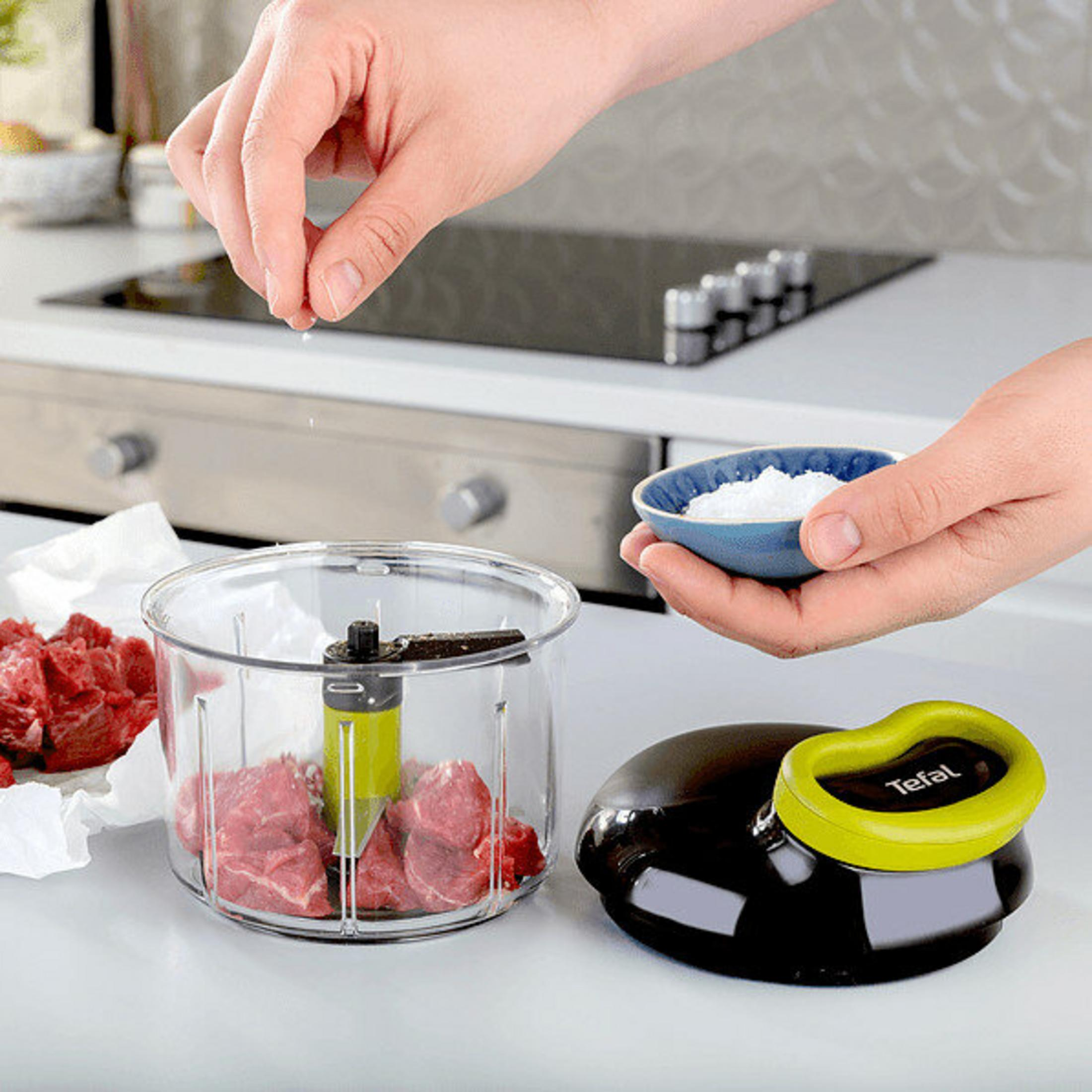 Osoba soli surowe mięso w przezroczystym rozdrabniaczu. Trzymana jest miska z solą, a w pobliżu rozdrabniacz i jego pokrywa.