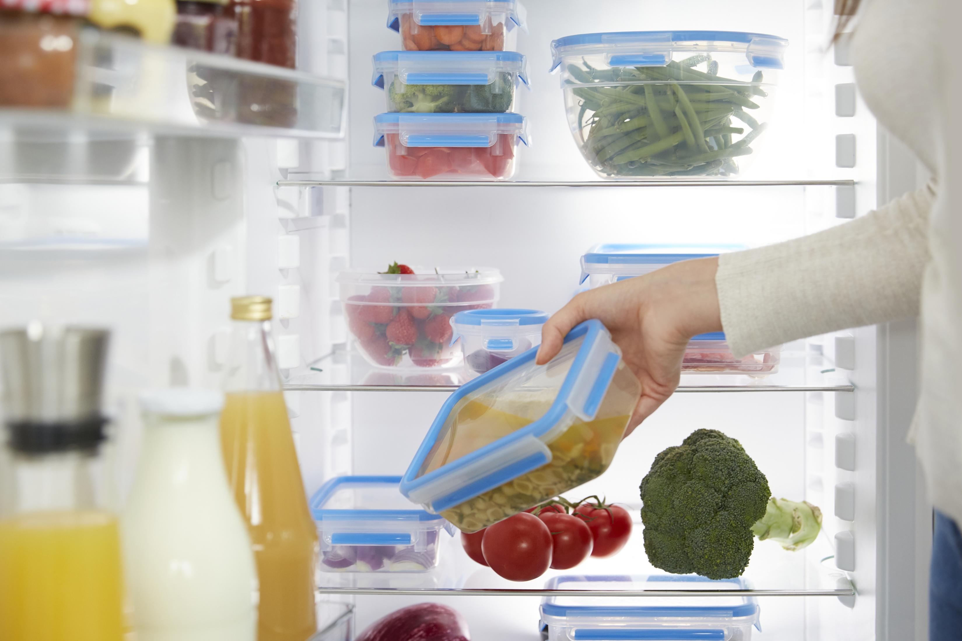 Una persona colocando un recipiente de comida en un refrigerador lleno de verduras y frutas.