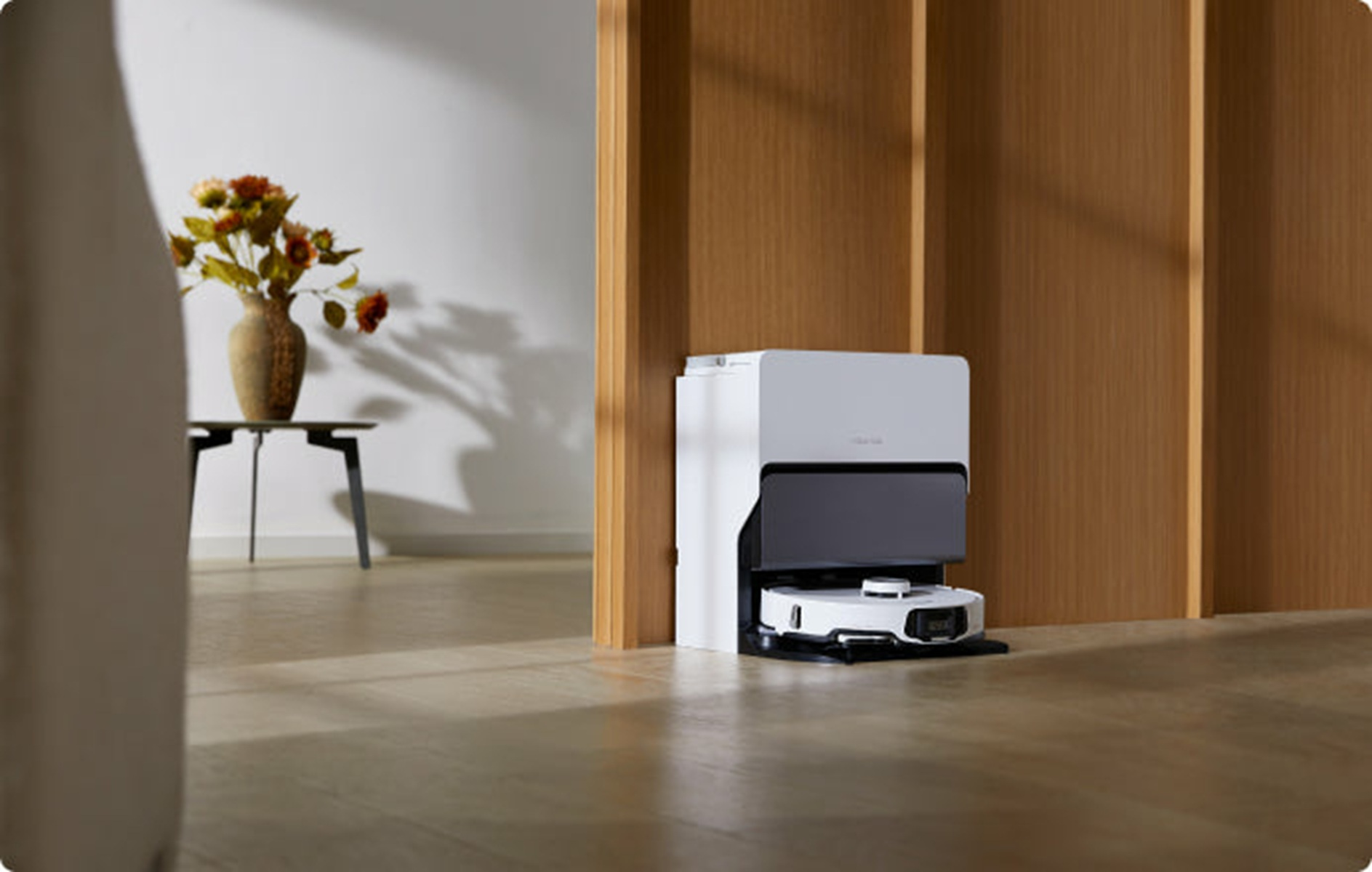 Robot odkurzający jest zadokowany w białej i czarnej stacji. Wazon z kwiatami stoi na stole. Drewniana rama drzwi.