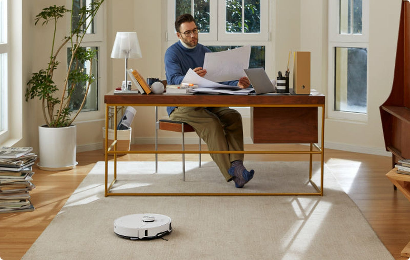 Mężczyzna w niebieskim swetrze siedzi przy biurku, przegląda papiery. Robot sprzątający na dywanie.