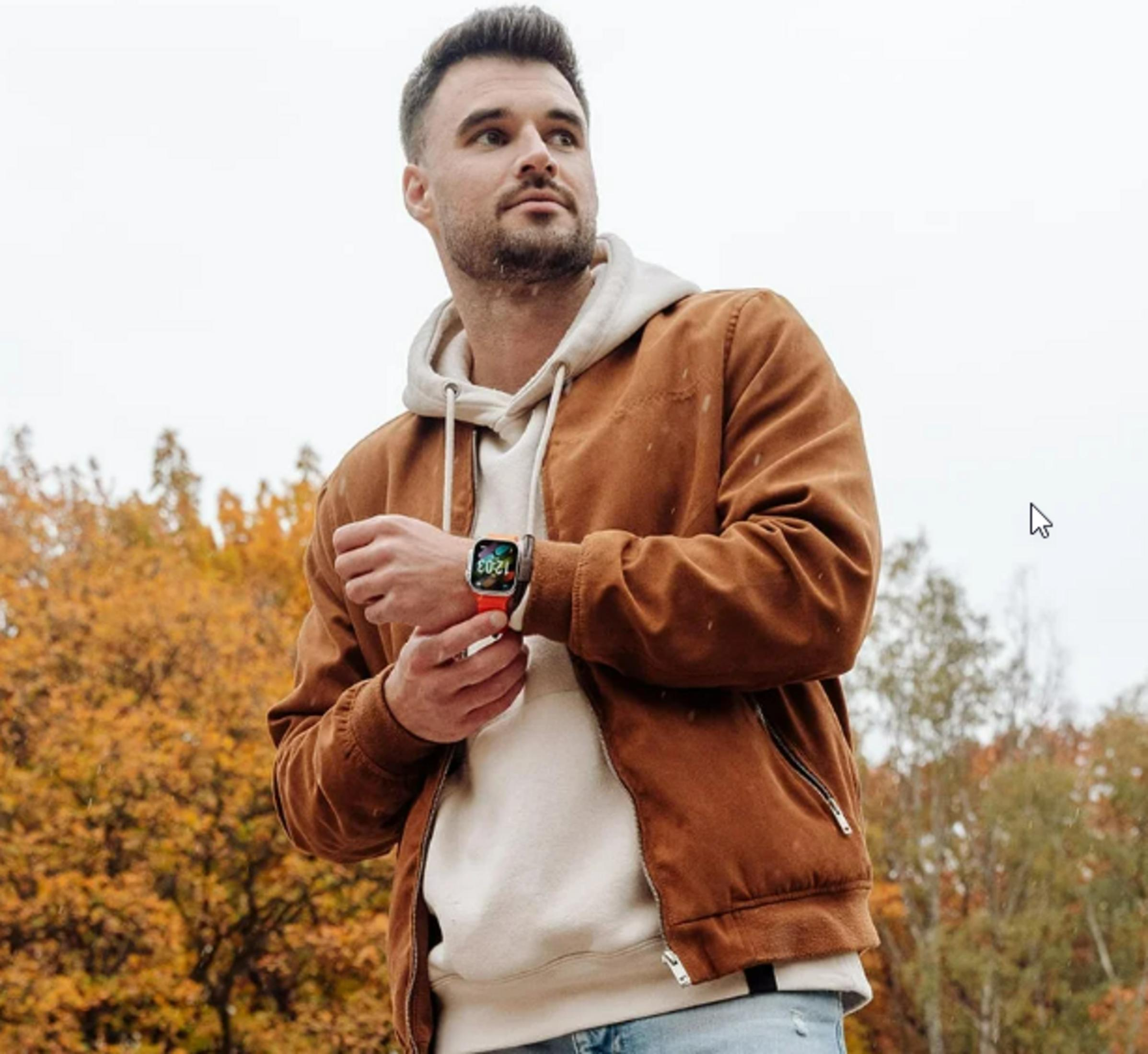 Mężczyzna na zewnątrz, w brązowej kurtce, białej bluzie z kapturem. Nosi smartwatch z czerwonym paskiem. Jesienne tło.