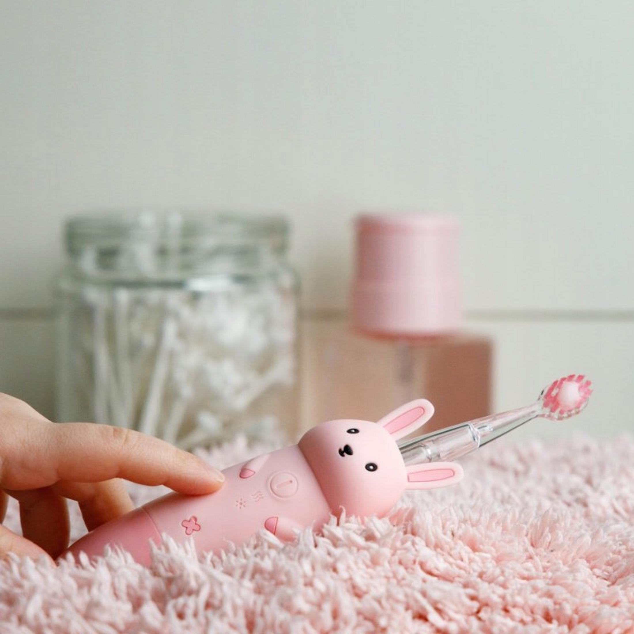 Uno spazzolino rosa a forma di coniglio è appoggiato su una superficie rosa e soffice. Una mano tiene lo spazzolino. Altri oggetti rosa e un barattolo sullo sfondo.
