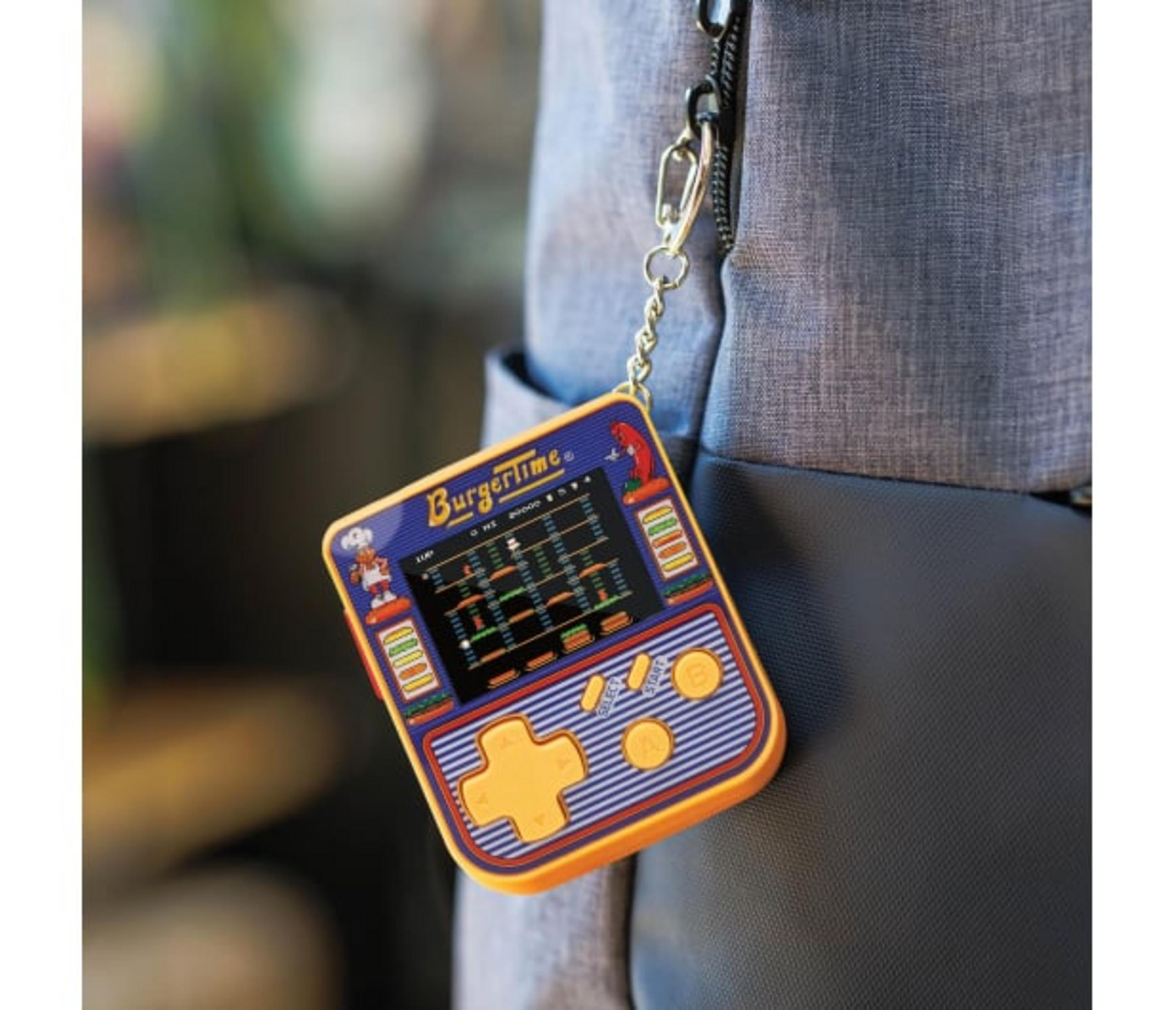 Przenośna gra BurgerTime jest przymocowana do torby. Jest pomarańczowa i niebieska, z ekranem gry. Tło jest rozmyte.