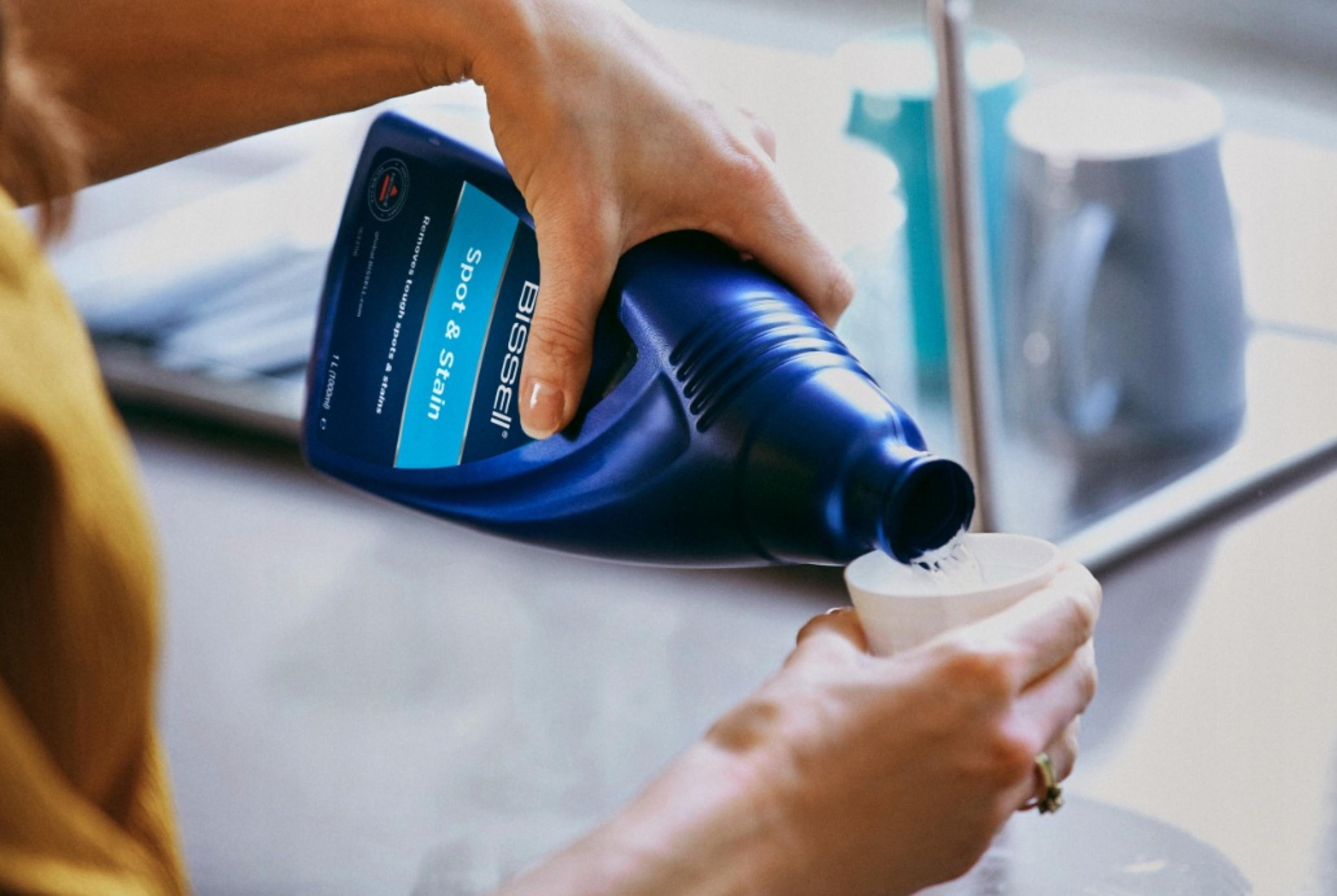 Eine Person gießt aus einer blauen Bissell-Flasche in einen weißen Becher. Die Flasche ist mit 'Spot & Stain' beschriftet.
