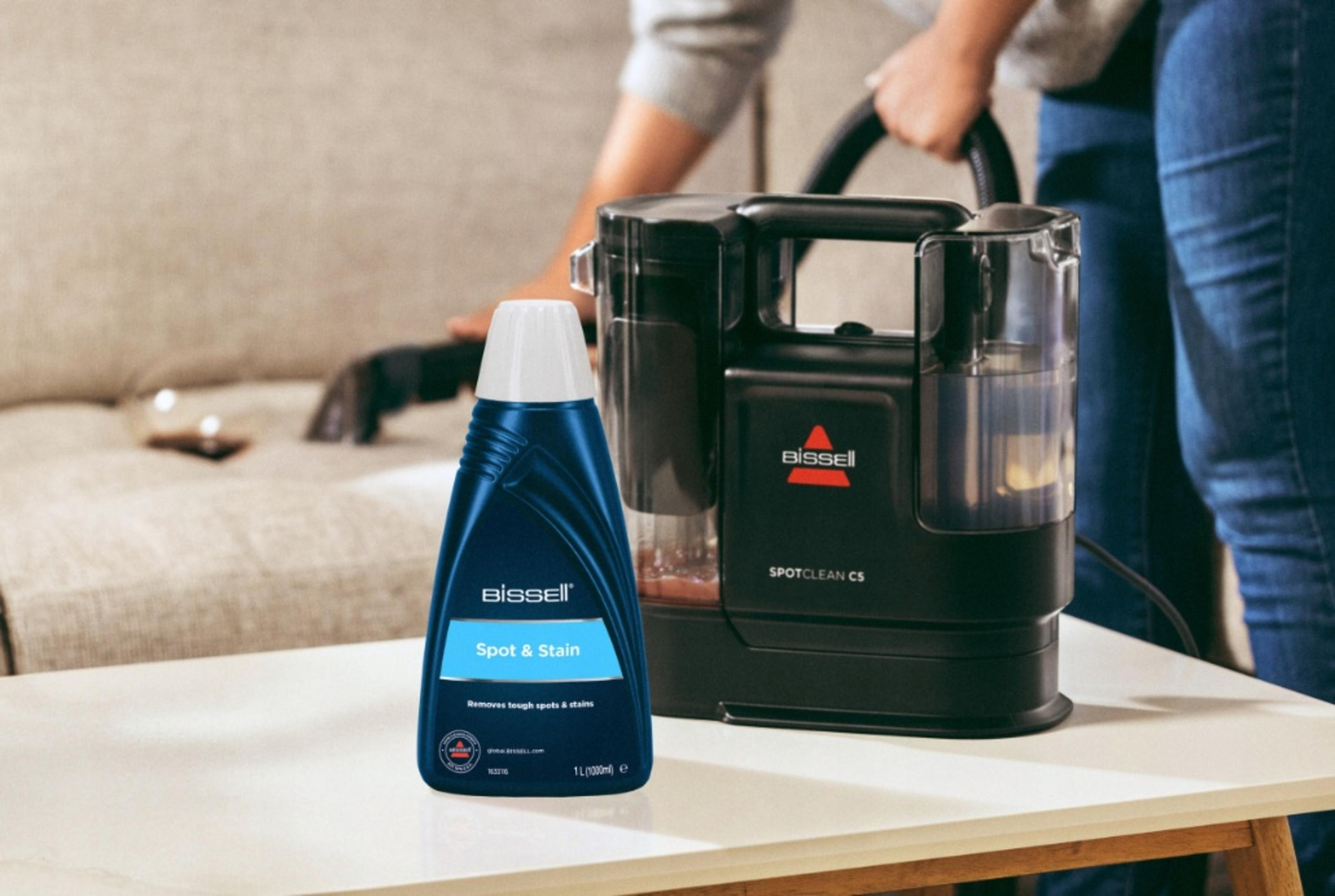 Eine Flasche Bissell Spot & Stain Reiniger und eine SpotClean C5 Maschine auf einem weißen Tisch, Person reinigt Sofa im Hintergrund.