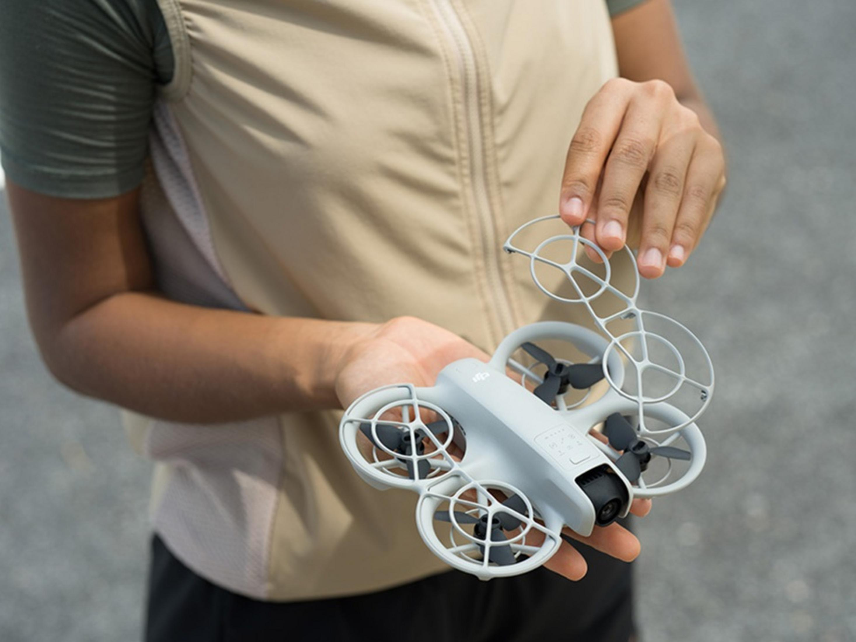 Una persona tiene in mano un drone grigio con protezioni. Lo sfondo è una superficie grigia sfocata.