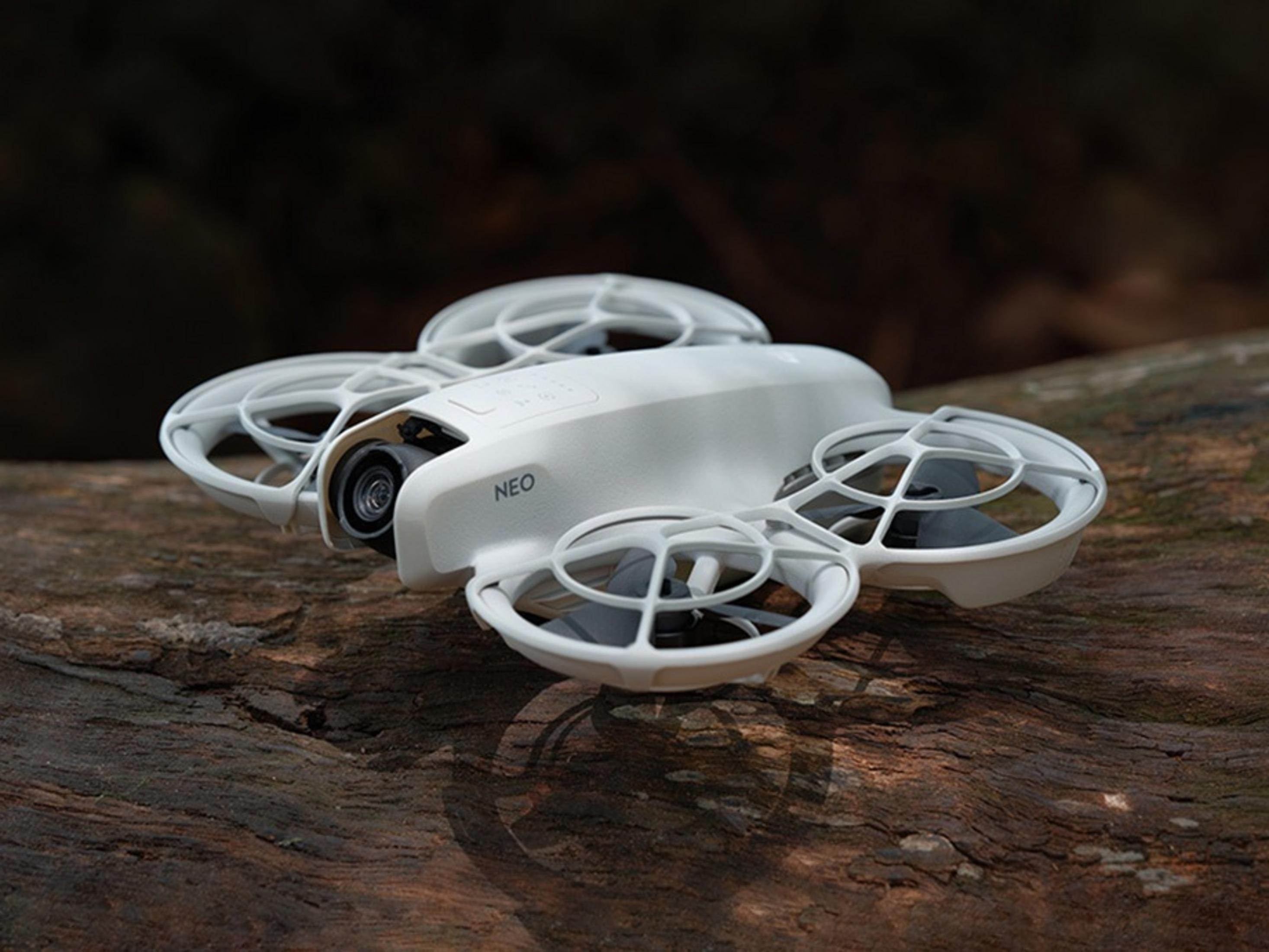 Un drone bianco con etichetta NEO poggia su un tronco marrone. Ha quattro eliche e una fotocamera. Sfondo scuro.