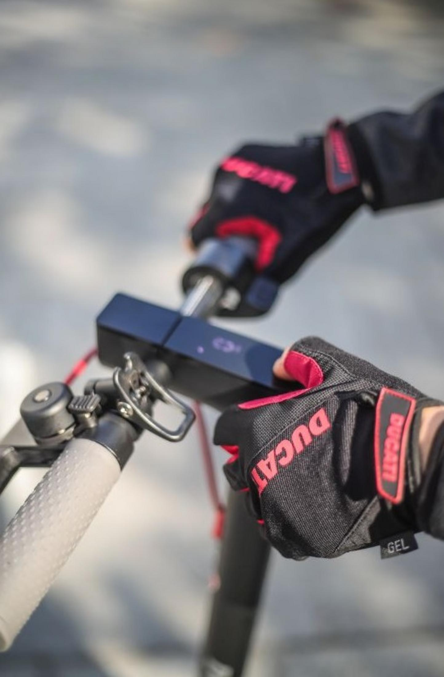 Mani con guanti Ducati neri che tengono il manubrio di un monopattino elettrico. I guanti hanno dettagli rossi e logo Ducati. Sfondo sfocato.