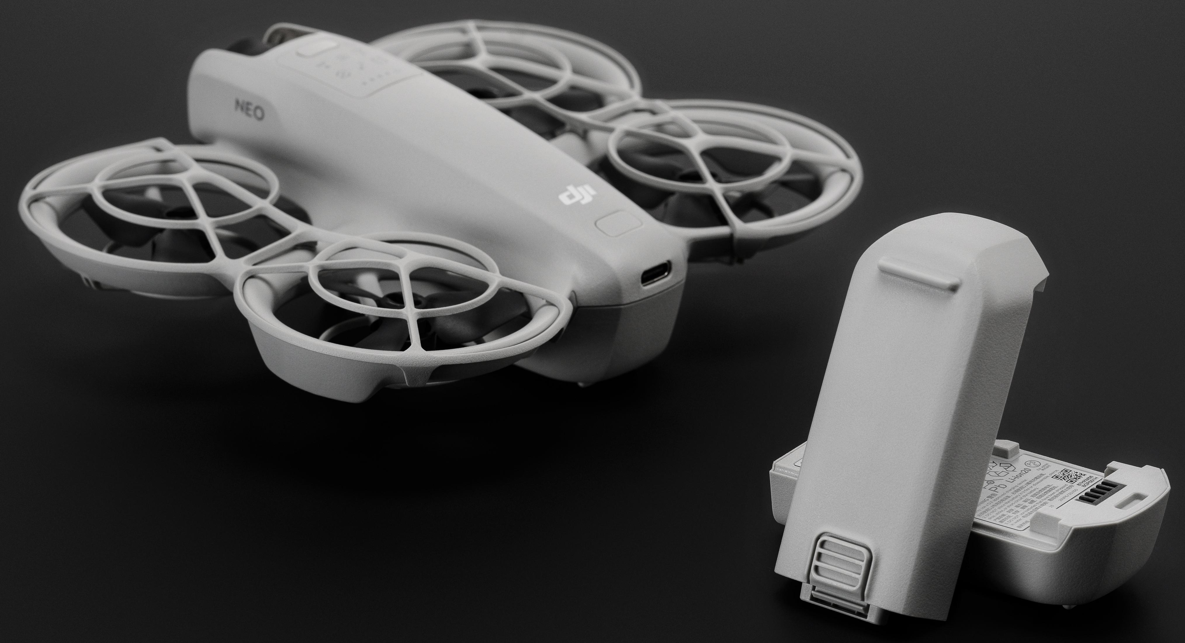 Un drone bianco e una batteria su una superficie nera. Il drone ha quattro eliche. La batteria è aperta.