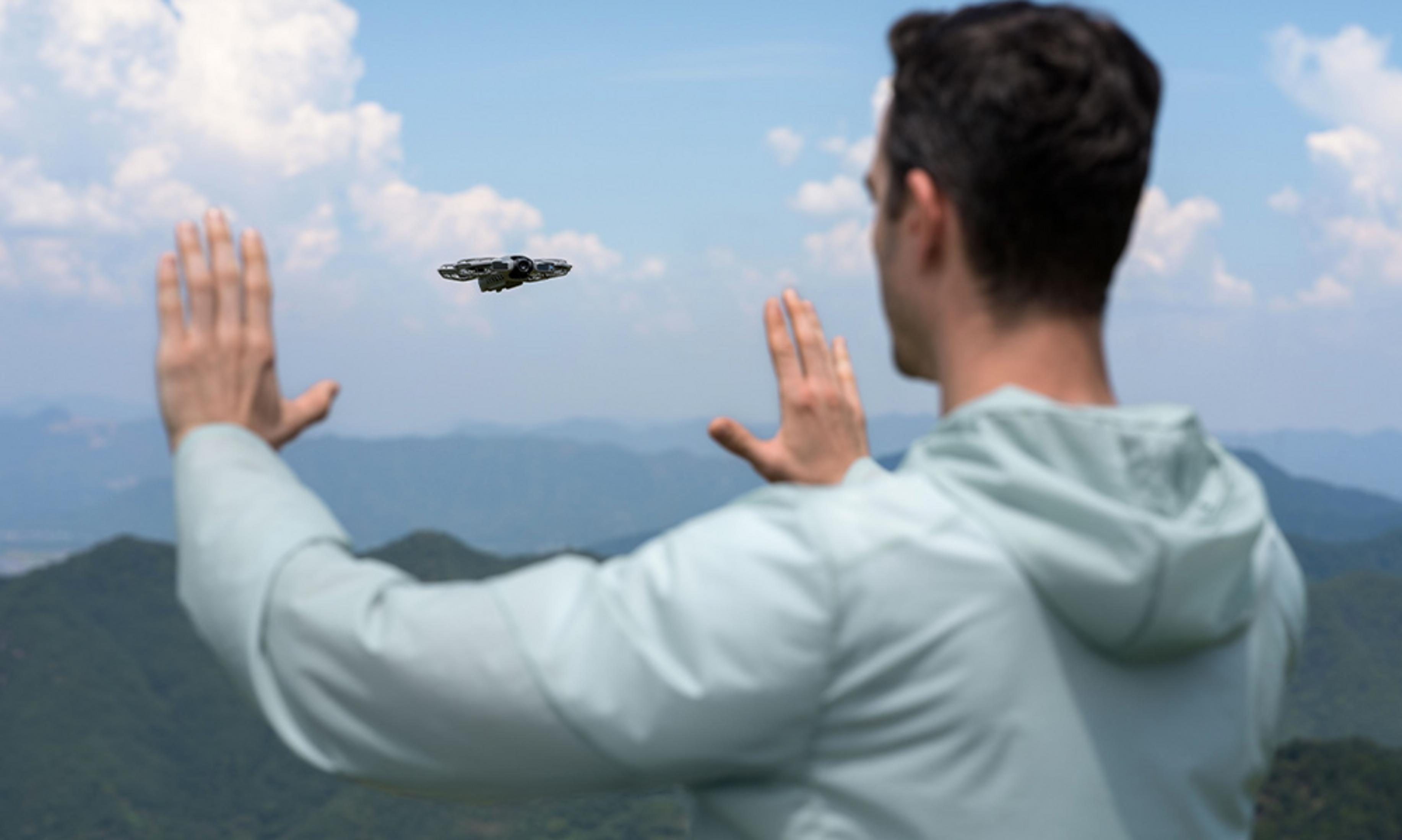 Mężczyzna z podniesionymi rękami, patrzy na drona na niebie. W tle góry. Pochmurne niebo.