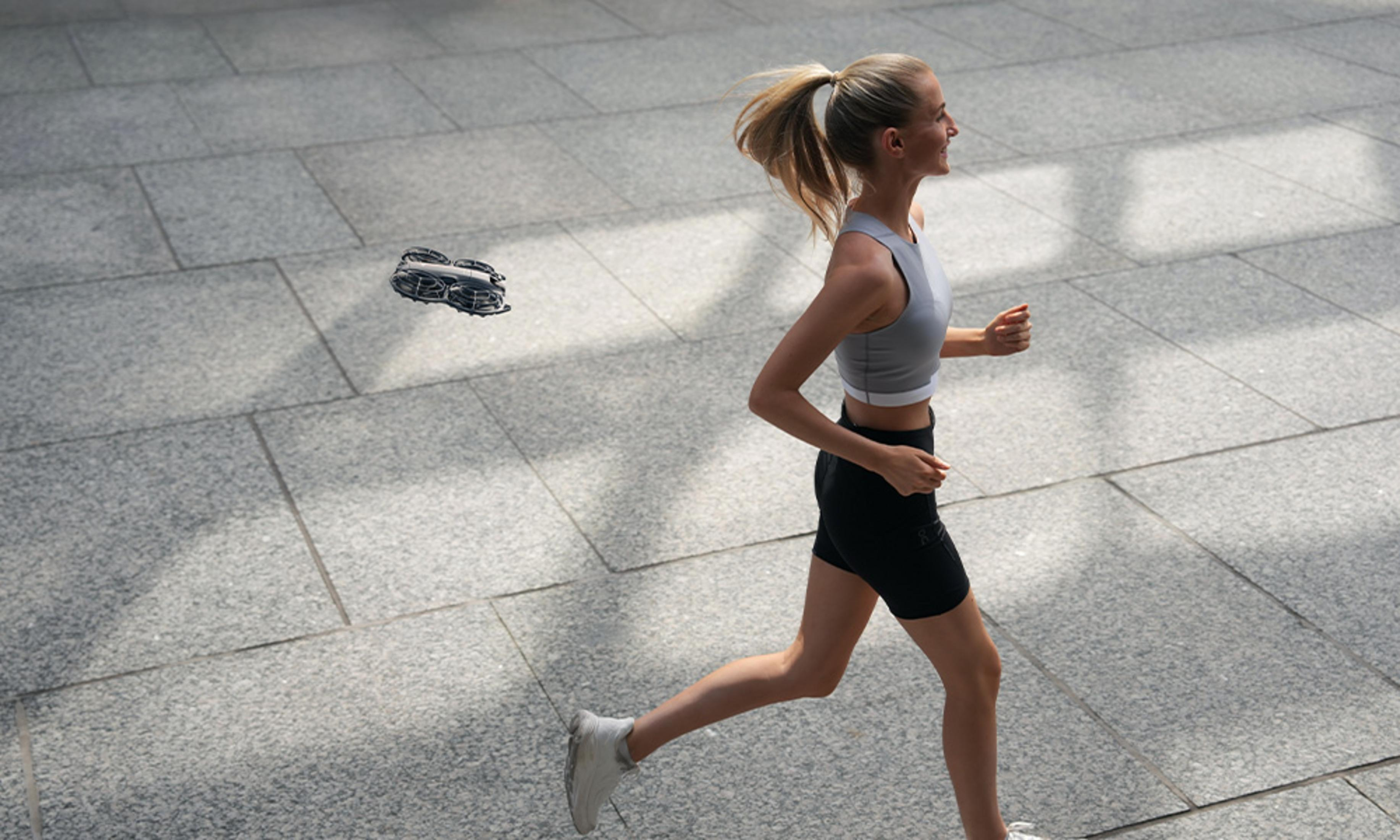 Kobieta biegnie po szarej, kafelkowanej powierzchni, obok unosi się dron. Ma na sobie strój sportowy.