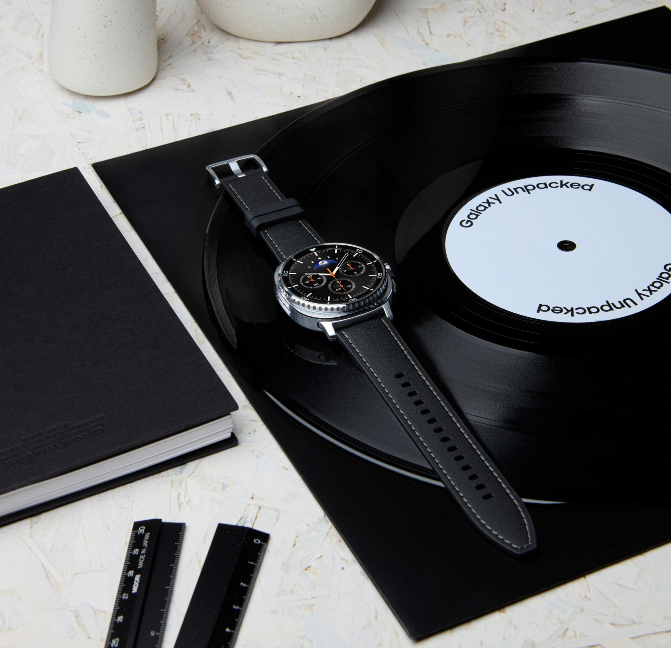 Un orologio su un vinile con la scritta 'Galaxy Unpacked'. Sono presenti anche un taccuino nero e dei righelli.