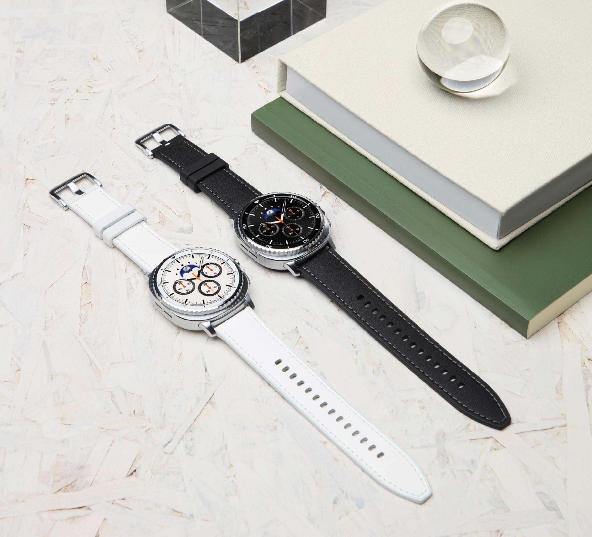 Due smartwatch con cinturini bianchi e neri su una superficie strutturata. Libri e una sfera di vetro nelle vicinanze.