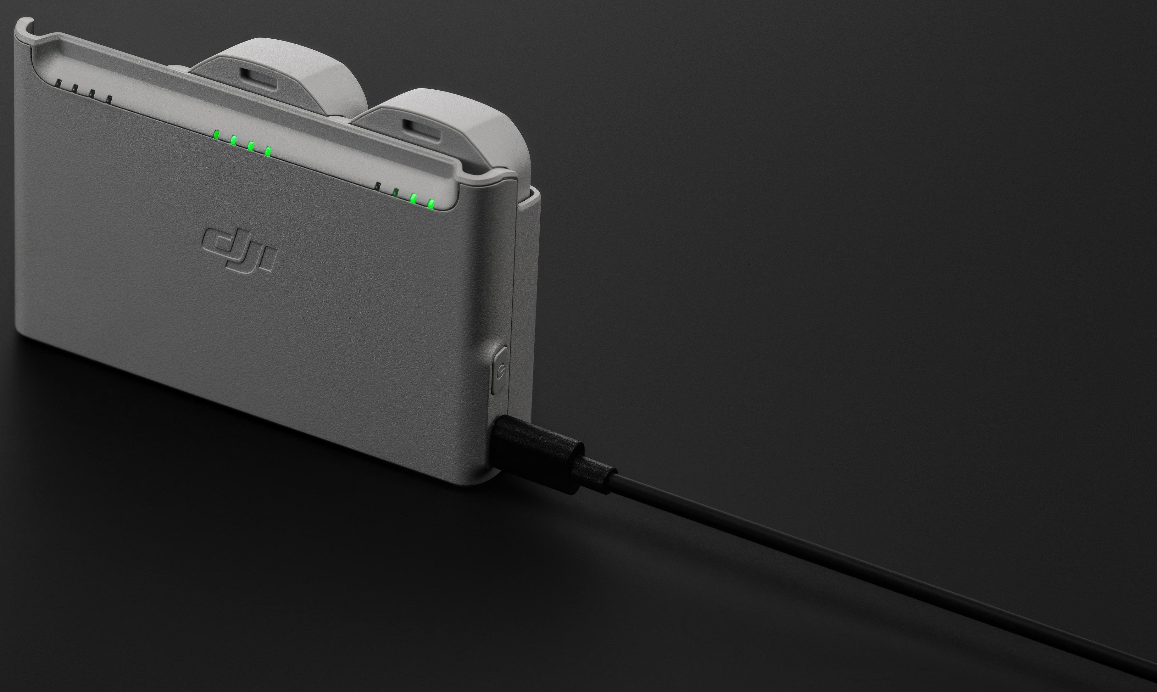 Un cargador de batería DJI gris con cable negro. Luces verdes indican estado de carga. El cargador está sobre una superficie negra.