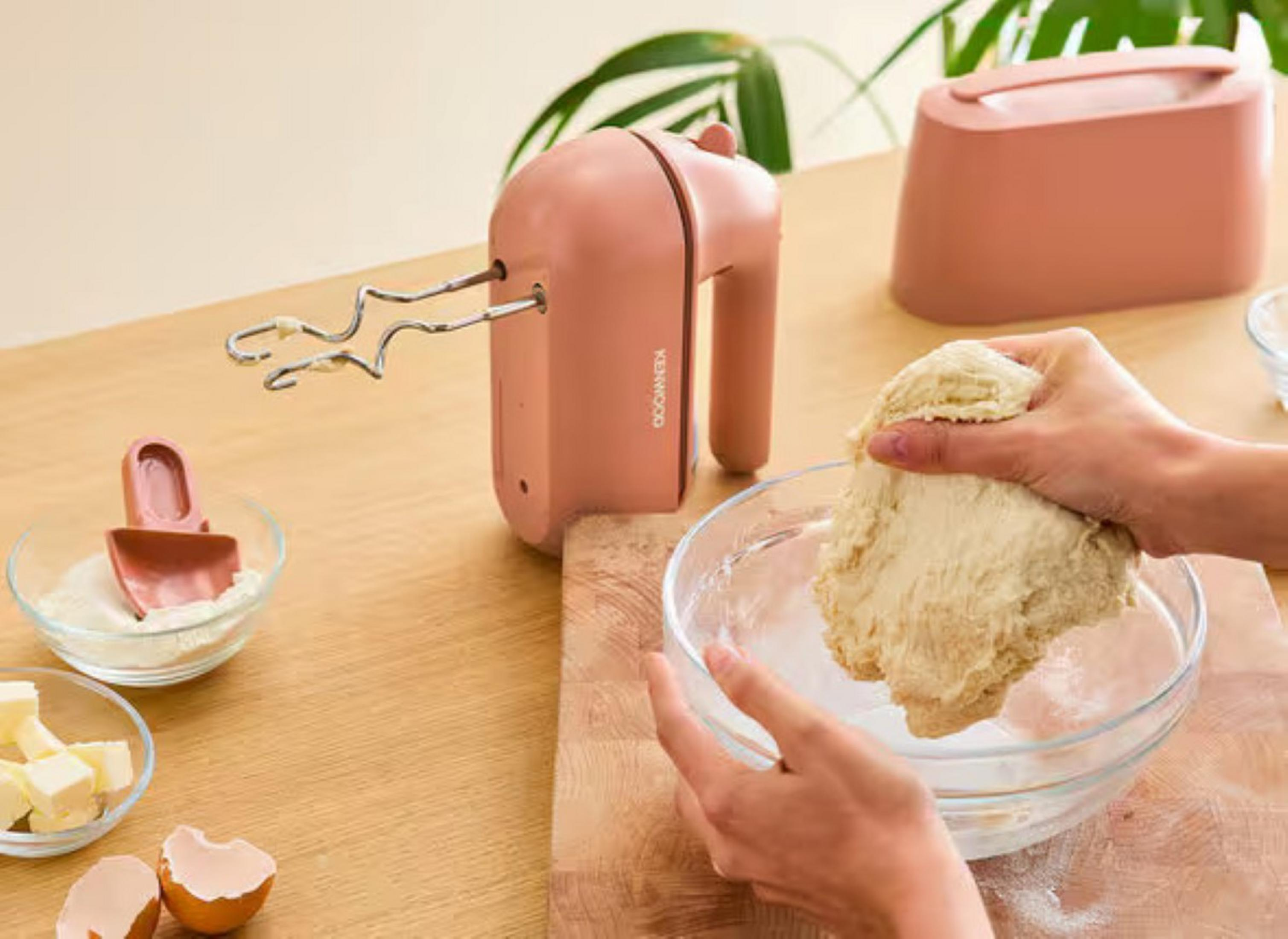 Ein rosa Mixer und Zutaten auf einem Holztisch. Hände kneten Teig in einer Glasschüssel.