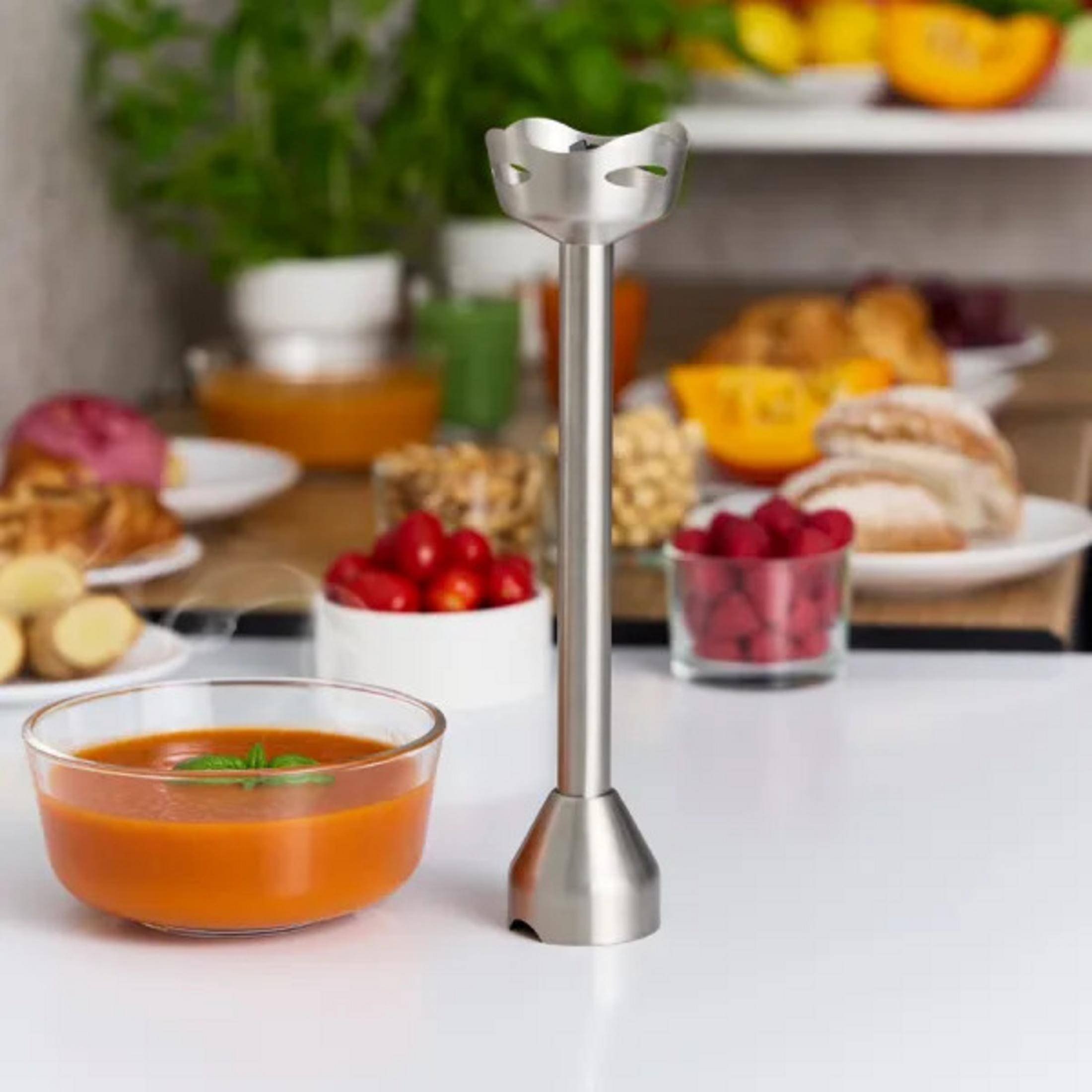 Un frullatore a immersione in metallo è su una superficie bianca. Una ciotola di zuppa, frutta e pane sullo sfondo.
