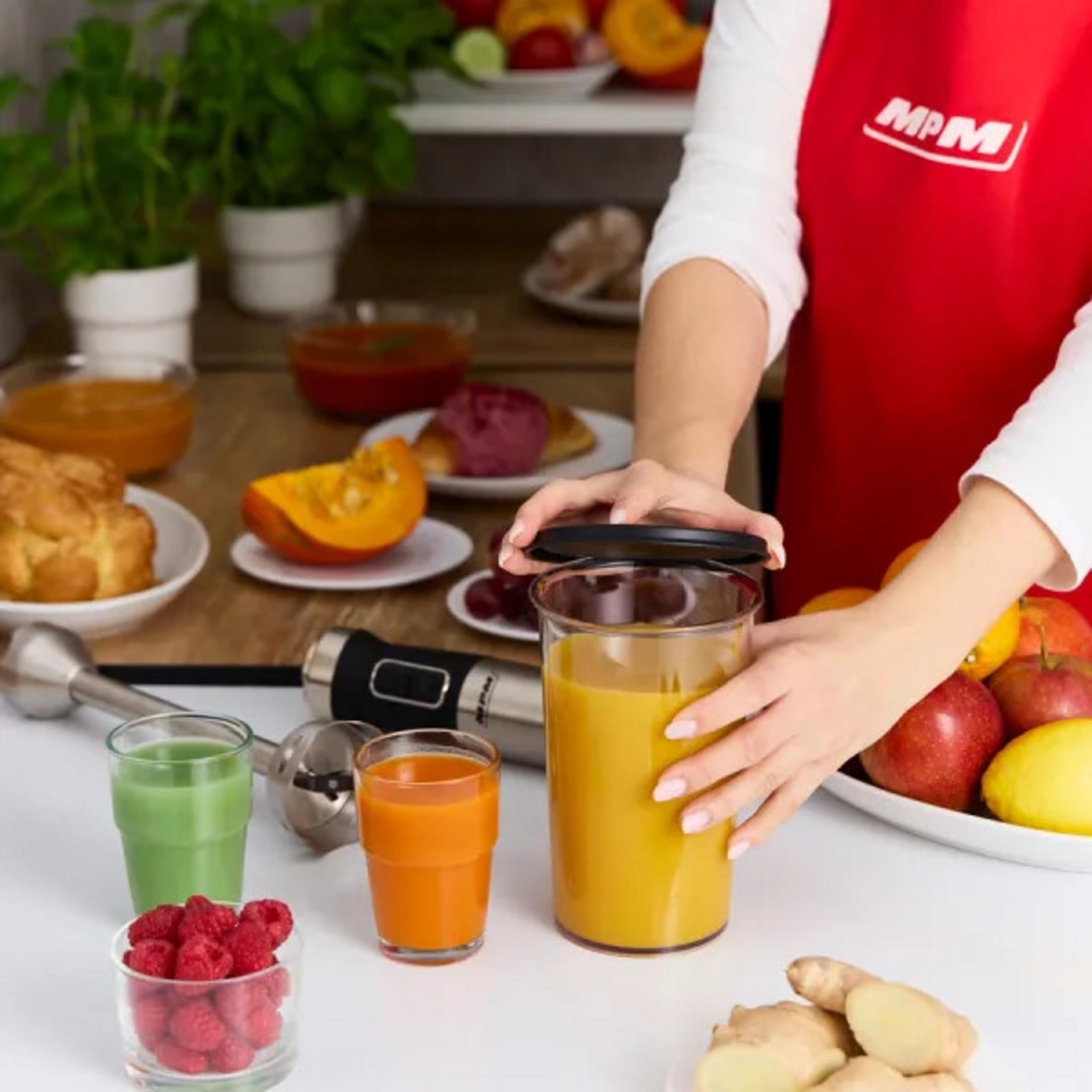 Una persona con un grembiule rosso prepara un frullato. Gli ingredienti includono frutta, verdura e un frullatore. Vari succhi sono in bicchieri.