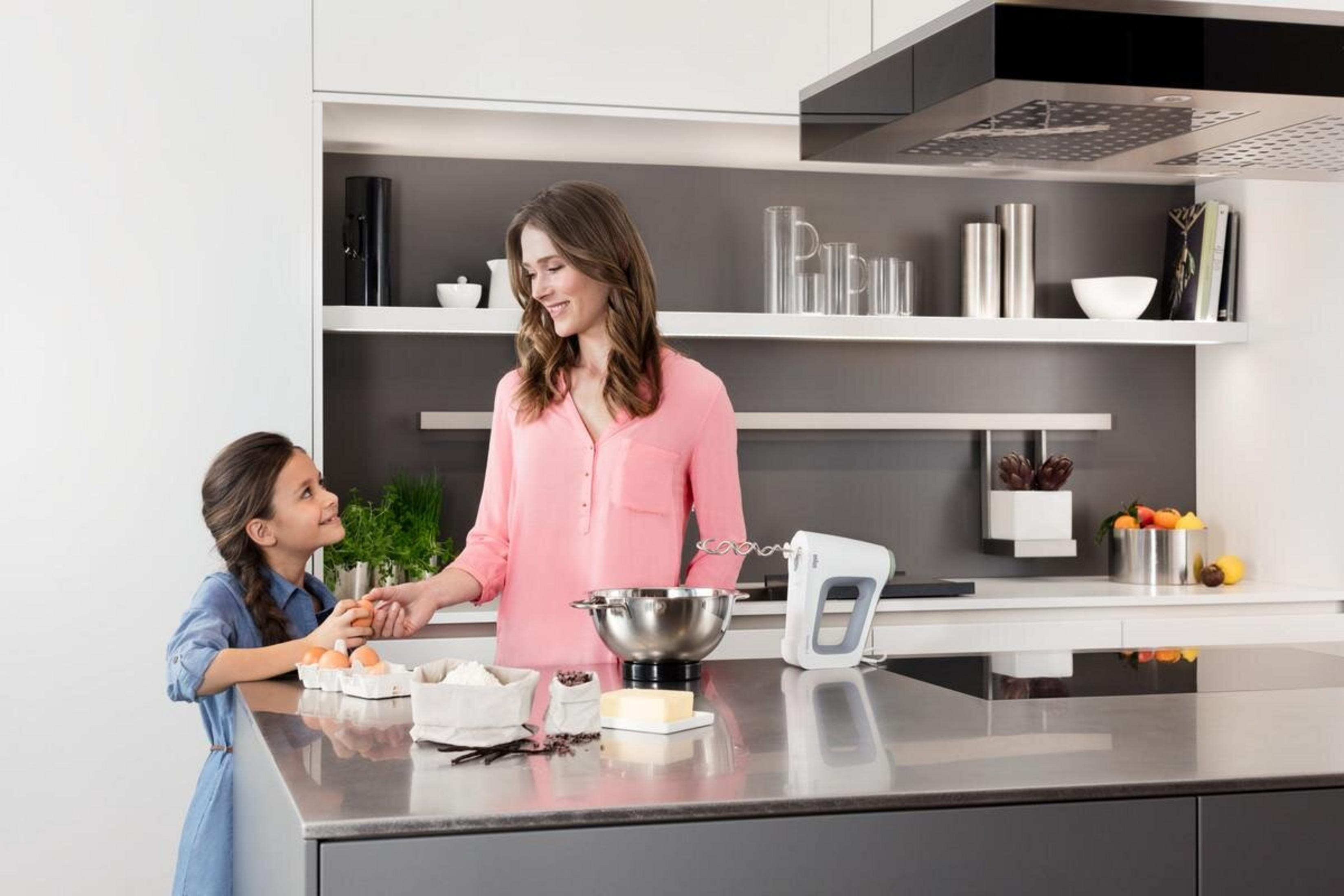 Kobieta i dziewczyna są w kuchni. Uśmiechają się. Składniki leżą na blacie. W pobliżu stoi mikser.