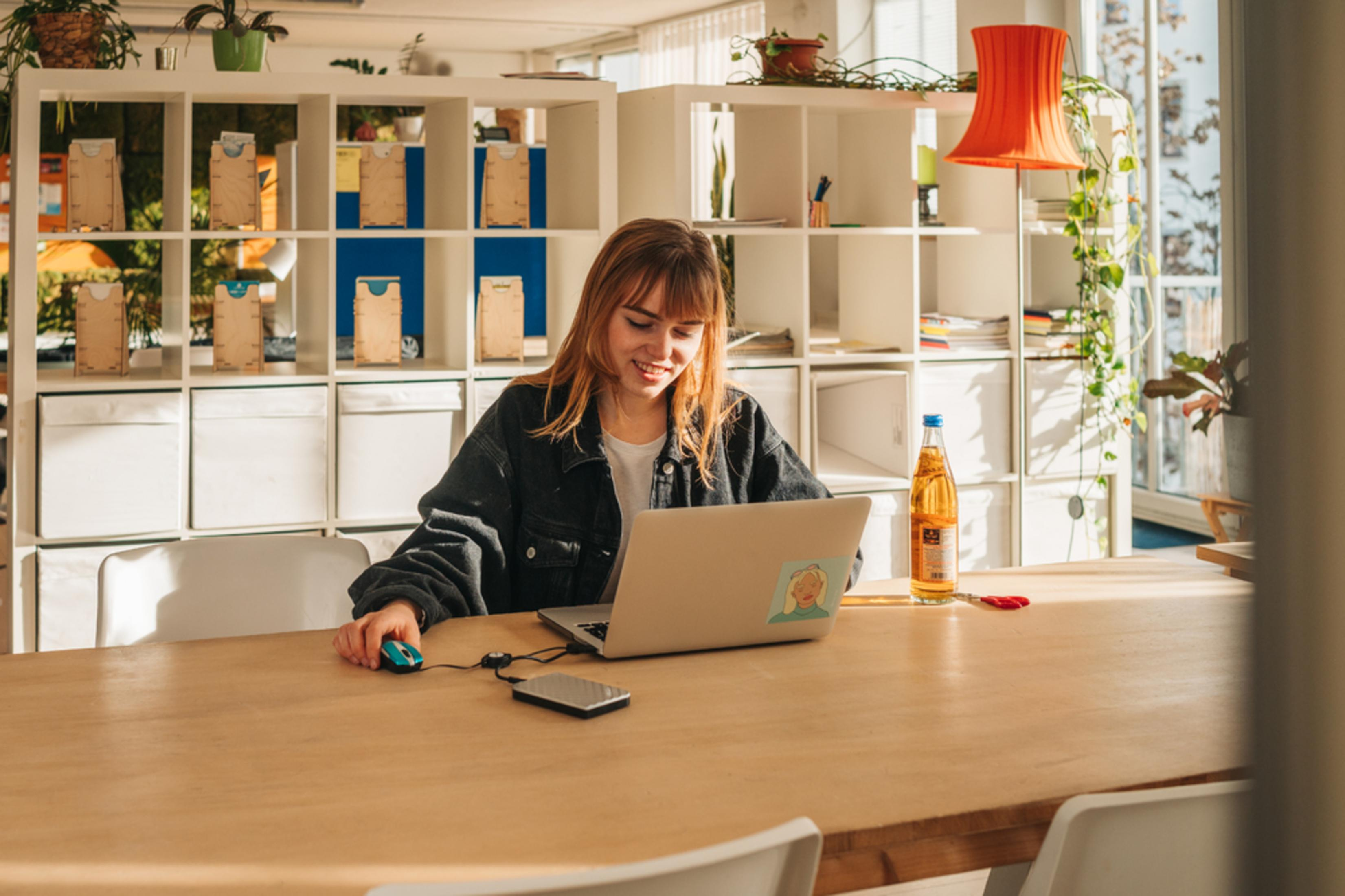 Kobieta używa laptopa, myszy i telefonu na drewnianym stole. Regały i okno w tle.