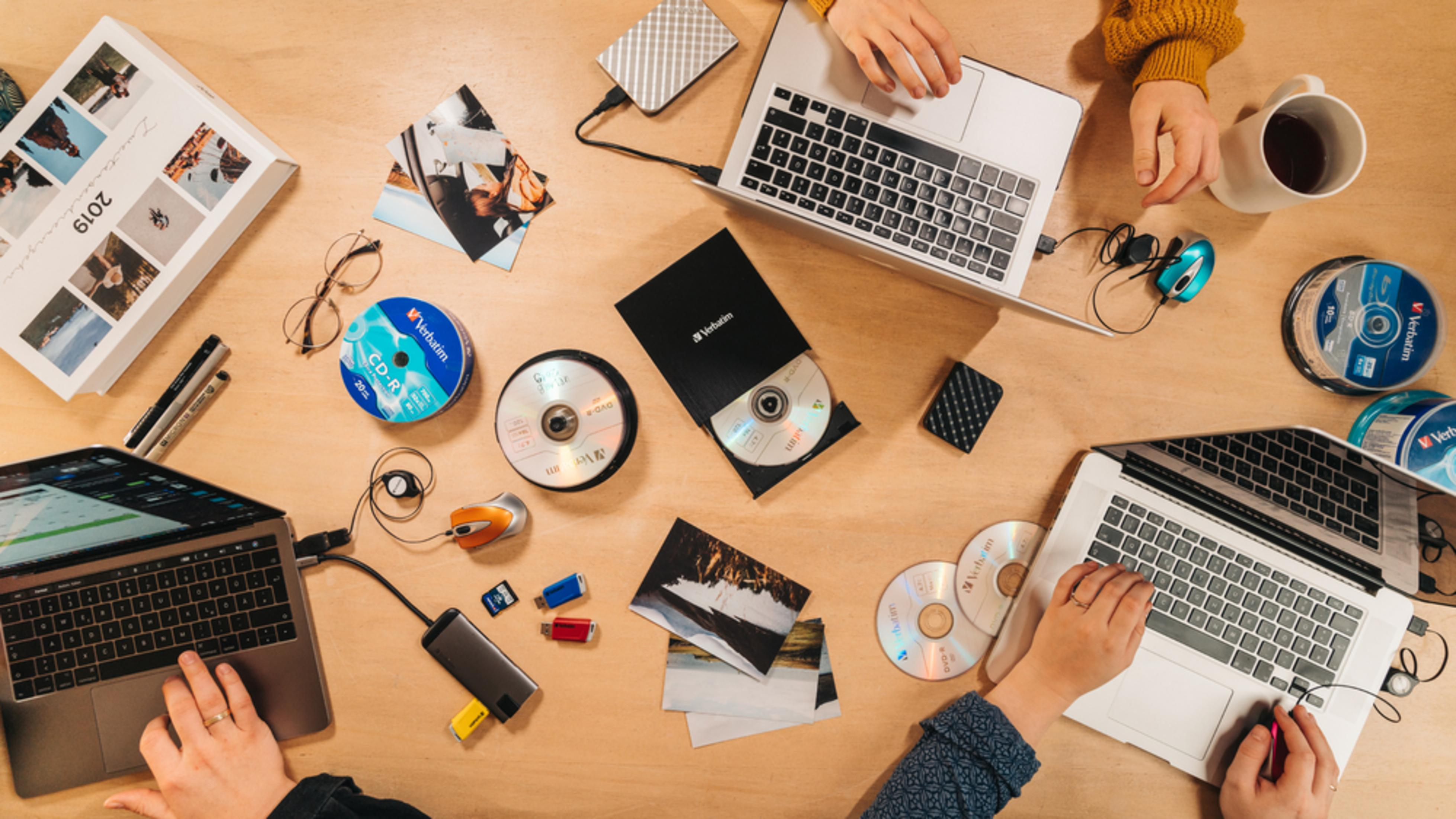 Drewniany stół z laptopami, płytami CD, zdjęciami i dłońmi. Ludzie pracują. Zawiera kubek, mysz i zewnętrzny dysk twardy.