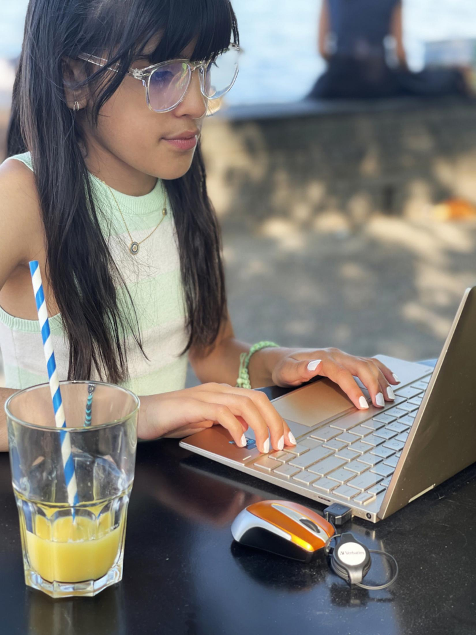A girl with glasses uses a laptop, a drink with a straw, a mouse and a headset on a table.