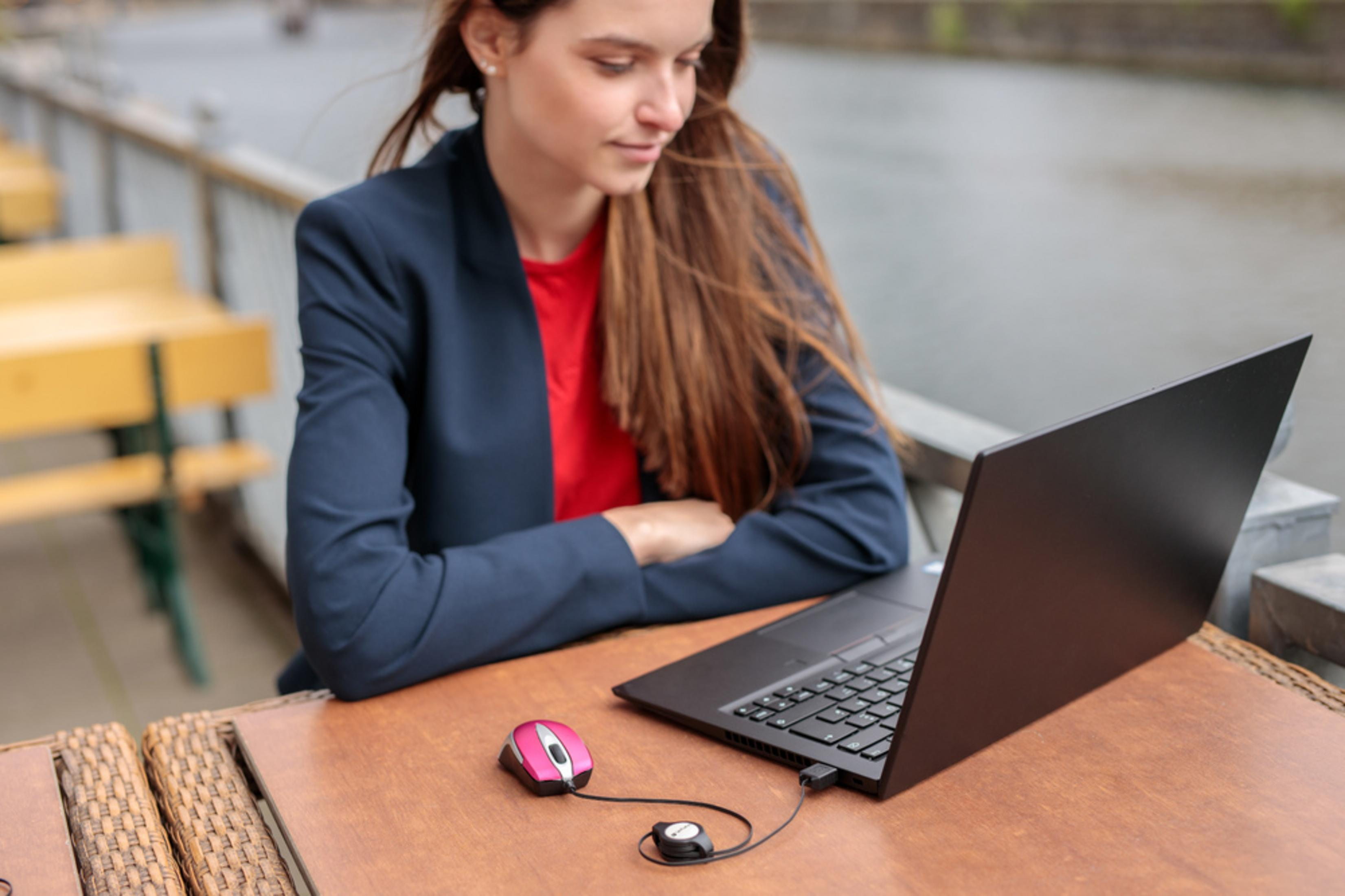 Kobieta w niebieskiej marynarce używa laptopa, różowa mysz na stole. Rzeka i most w tle.