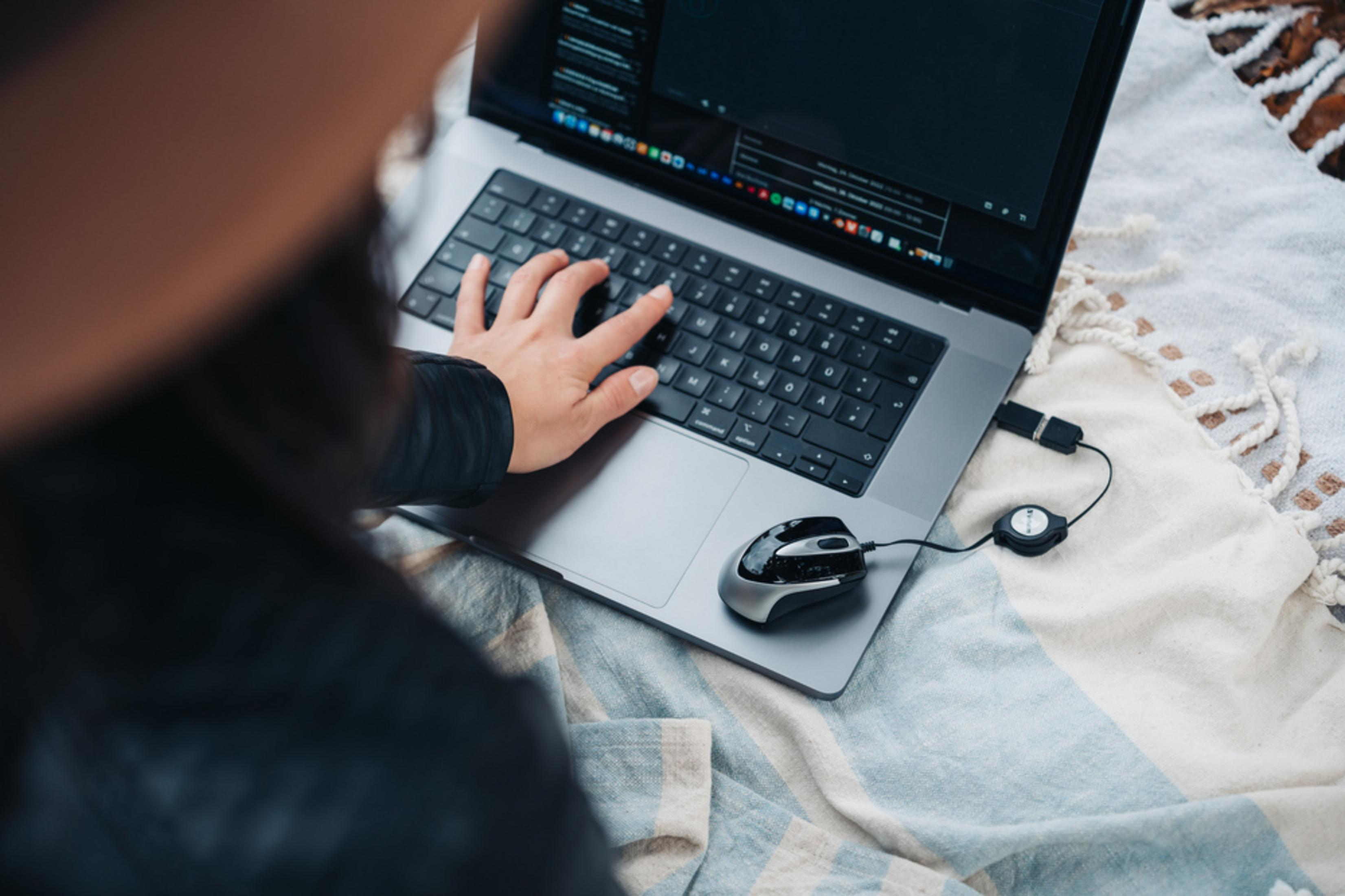 Eine Person benutzt einen Laptop im Freien. Eine Hand tippt auf der Tastatur. Eine Maus ist angeschlossen. Der Hintergrund ist eine Decke.