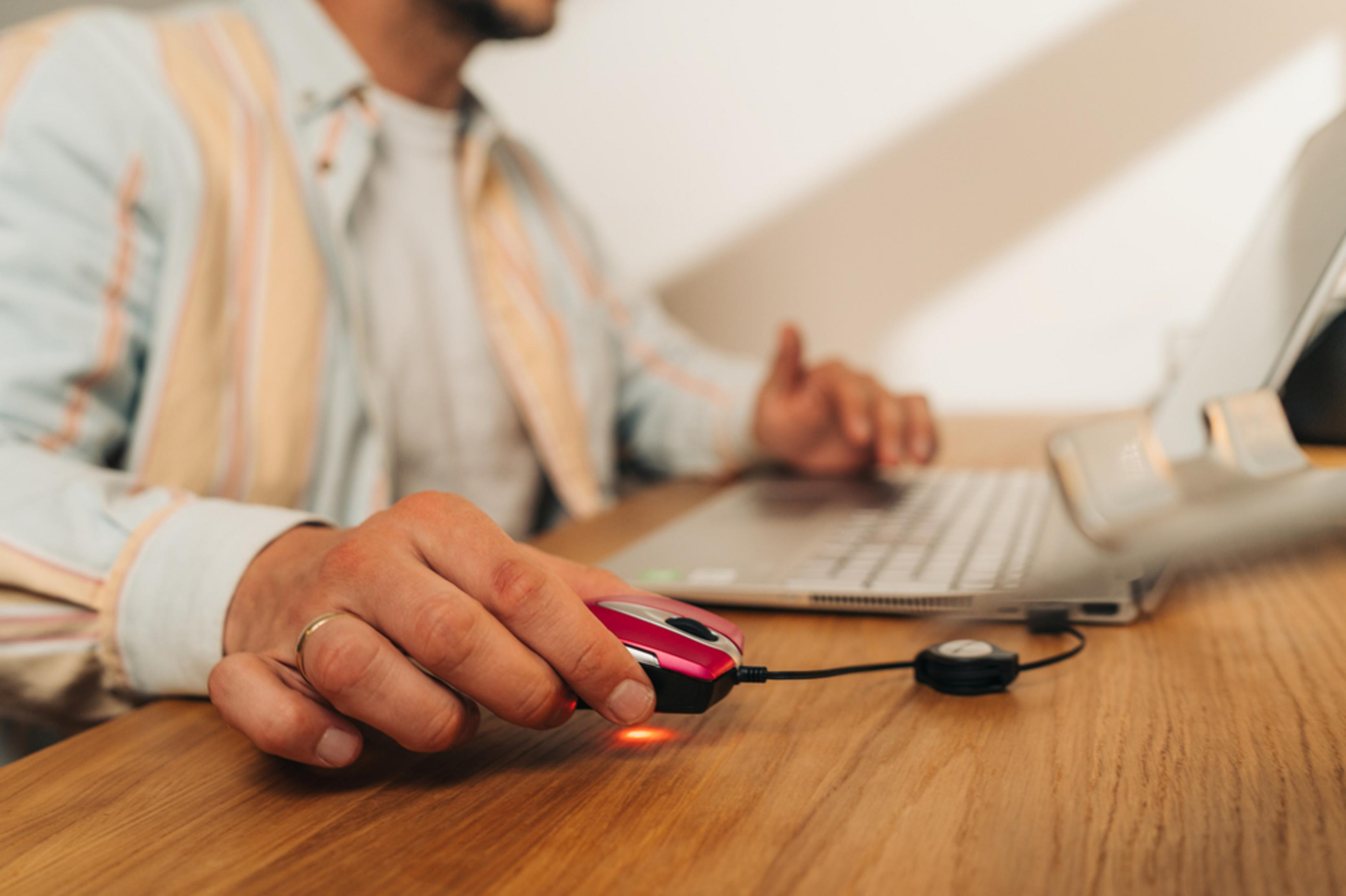 Osoba używająca różowej myszy podłączonej do laptopa na drewnianym biurku. Pierścionek na palcu.