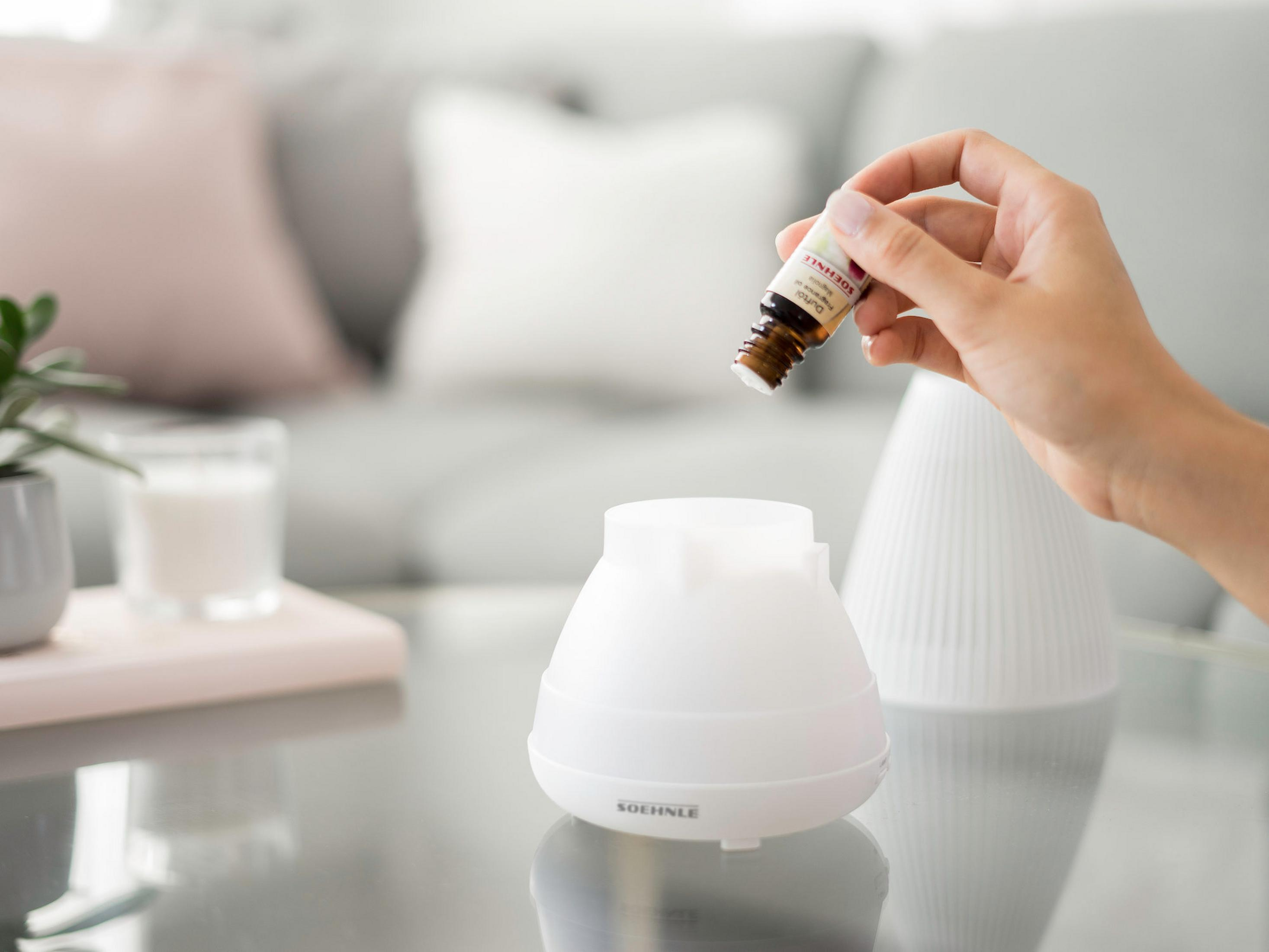 Una mano versa olio da una bottiglia in un diffusore bianco. Un divano e altri oggetti sono sullo sfondo.