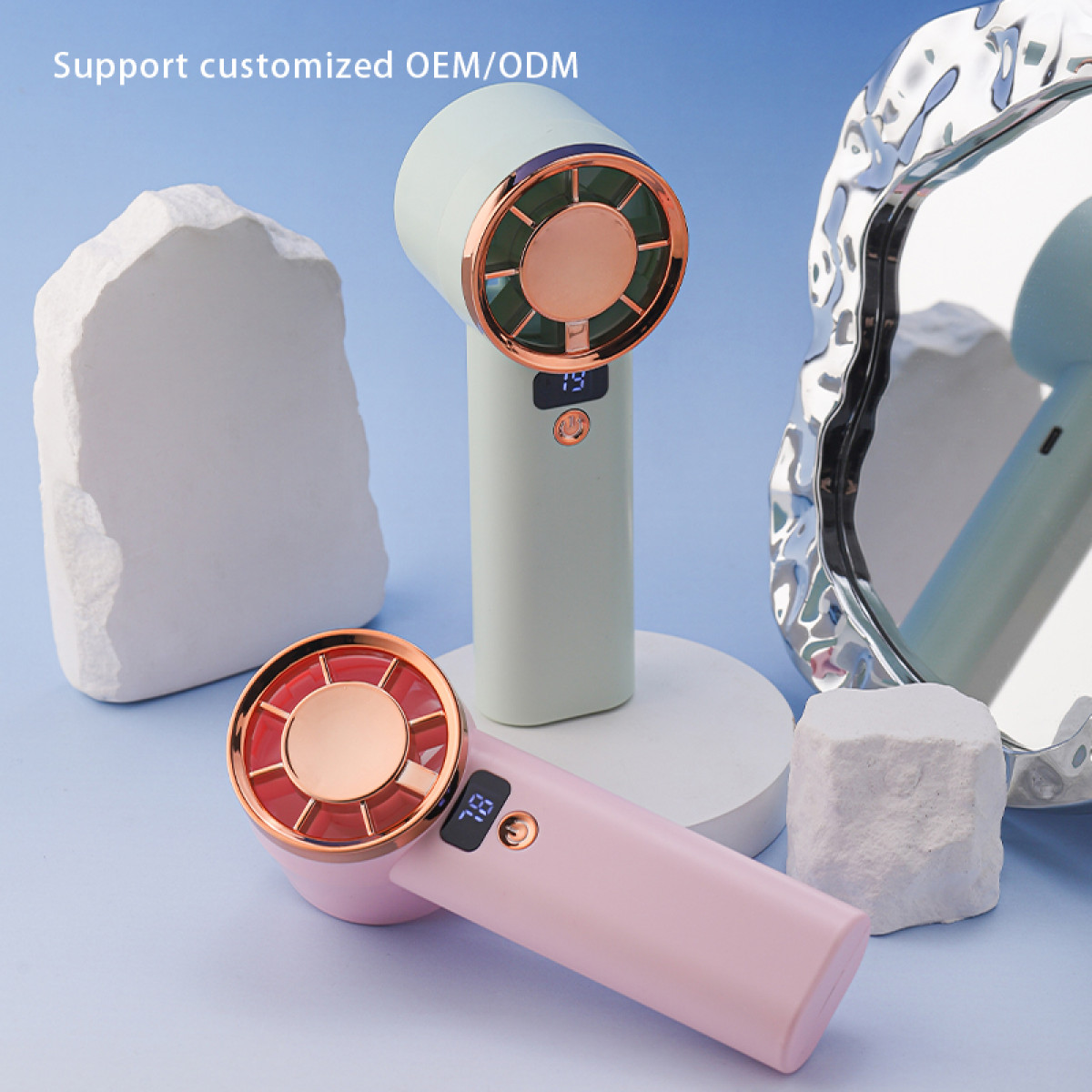 Dwa wentylatory ręczne, jeden różowy i jeden miętowy, na białych platformach na niebieskim tle, z lustrem.