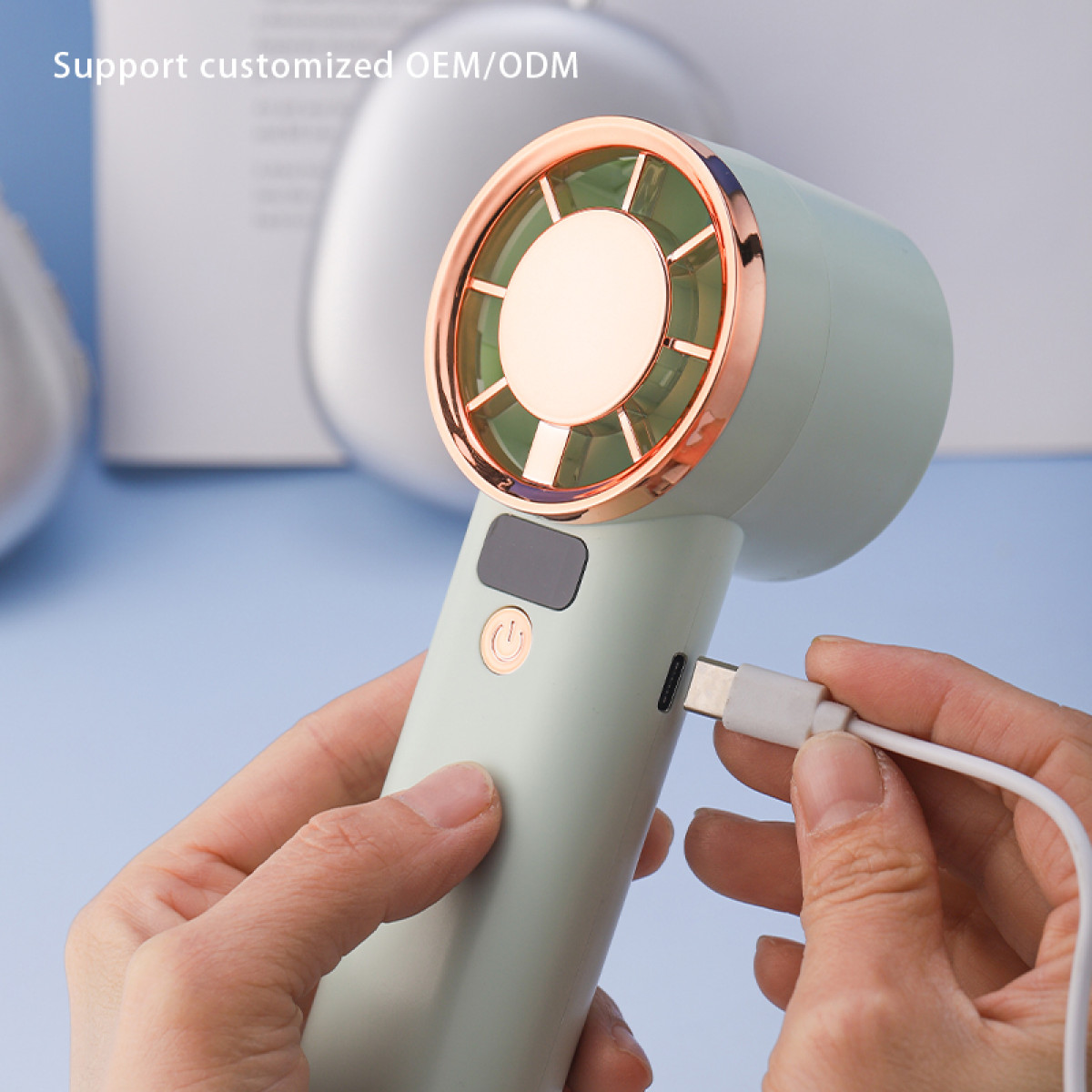 Wentylator ręczny w kolorze miętowej zieleni ze złotymi akcentami jest ładowany białym kablem. Tło jest rozmyte.