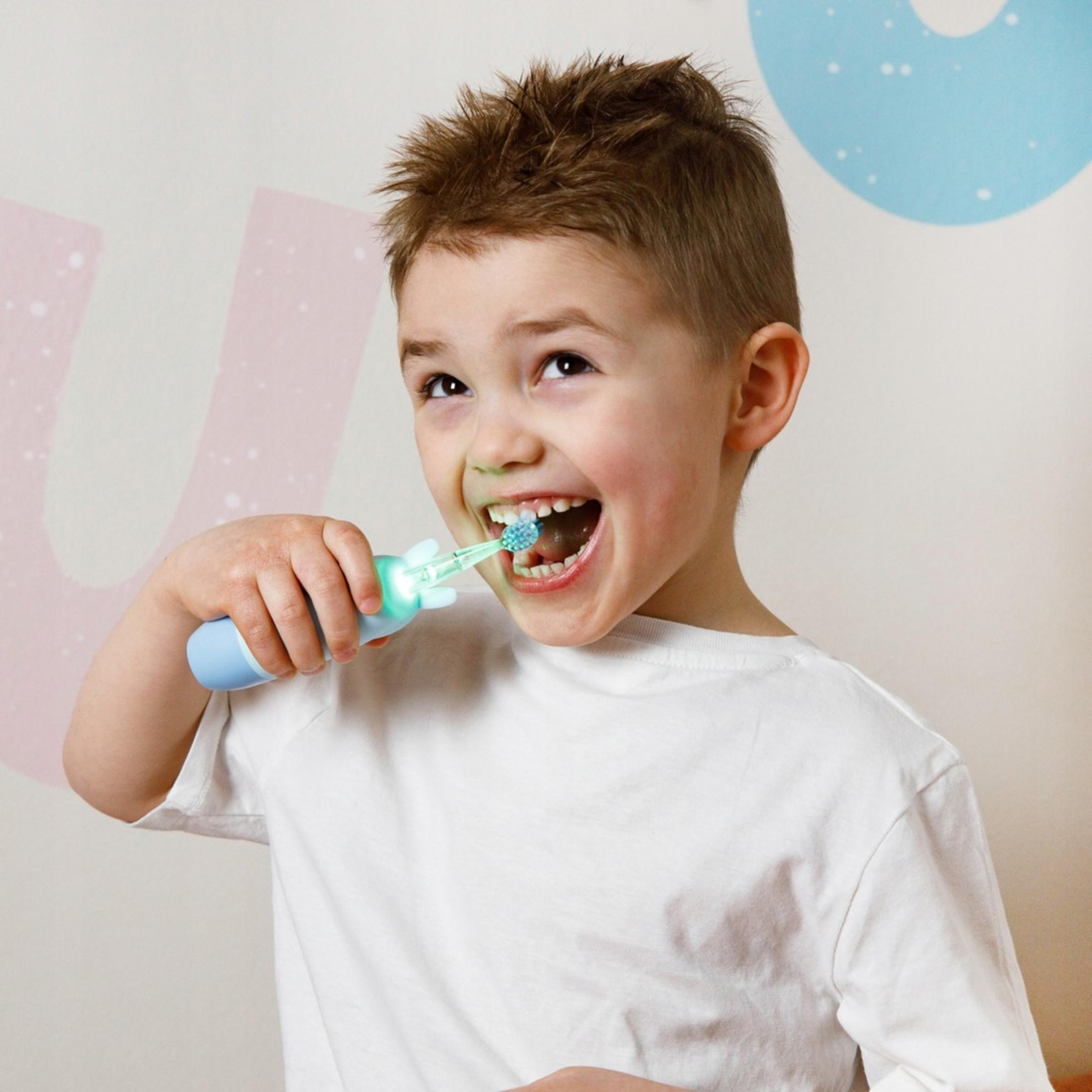 Un niño se cepilla los dientes con un cepillo de dientes azul y verde, sonriendo. El fondo tiene formas rosas y azules.