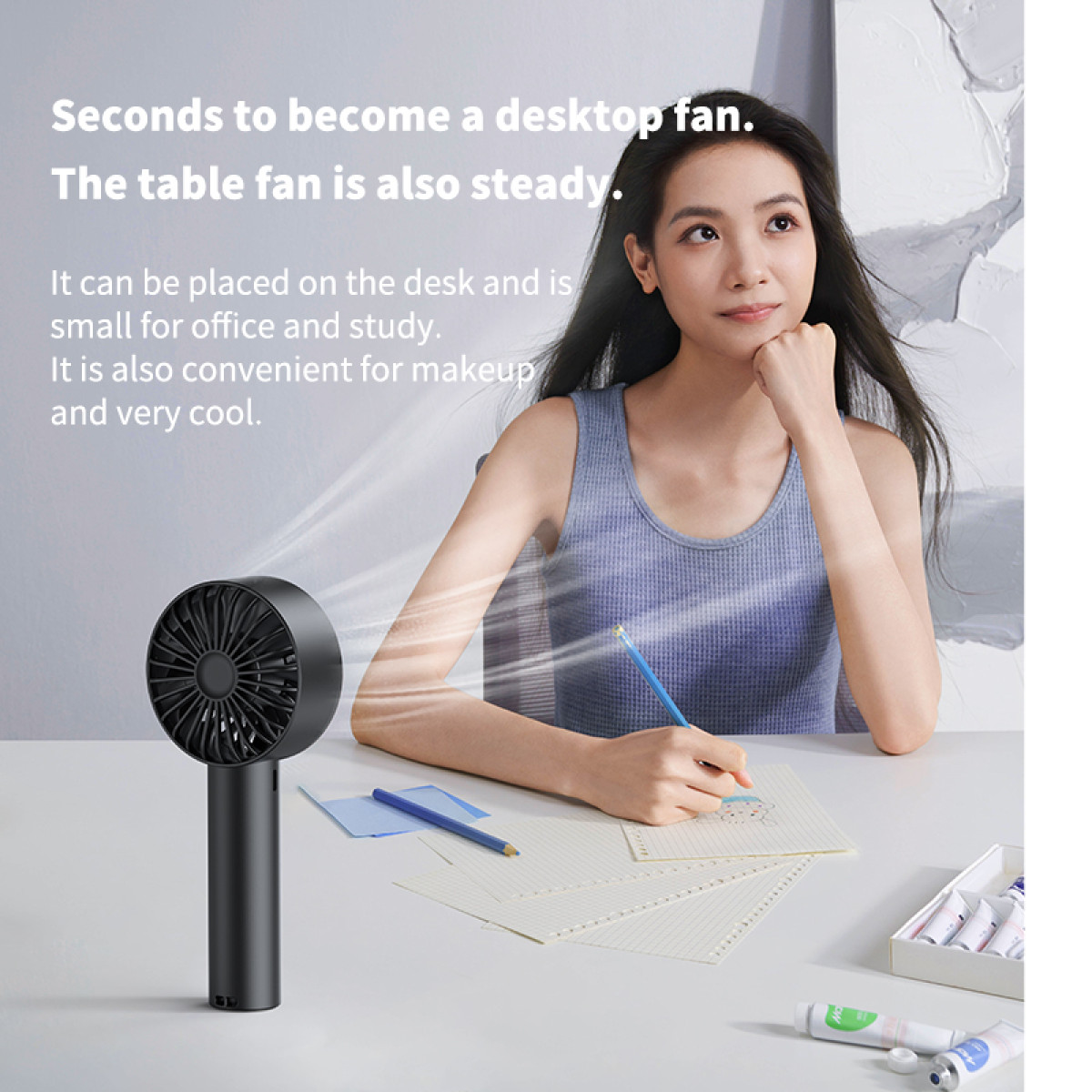 Kobieta siedzi przy biurku, patrząc w górę, z wentylatorem na pierwszym planie. Na biurku leżą papiery, ołówki i tubki do makijażu.