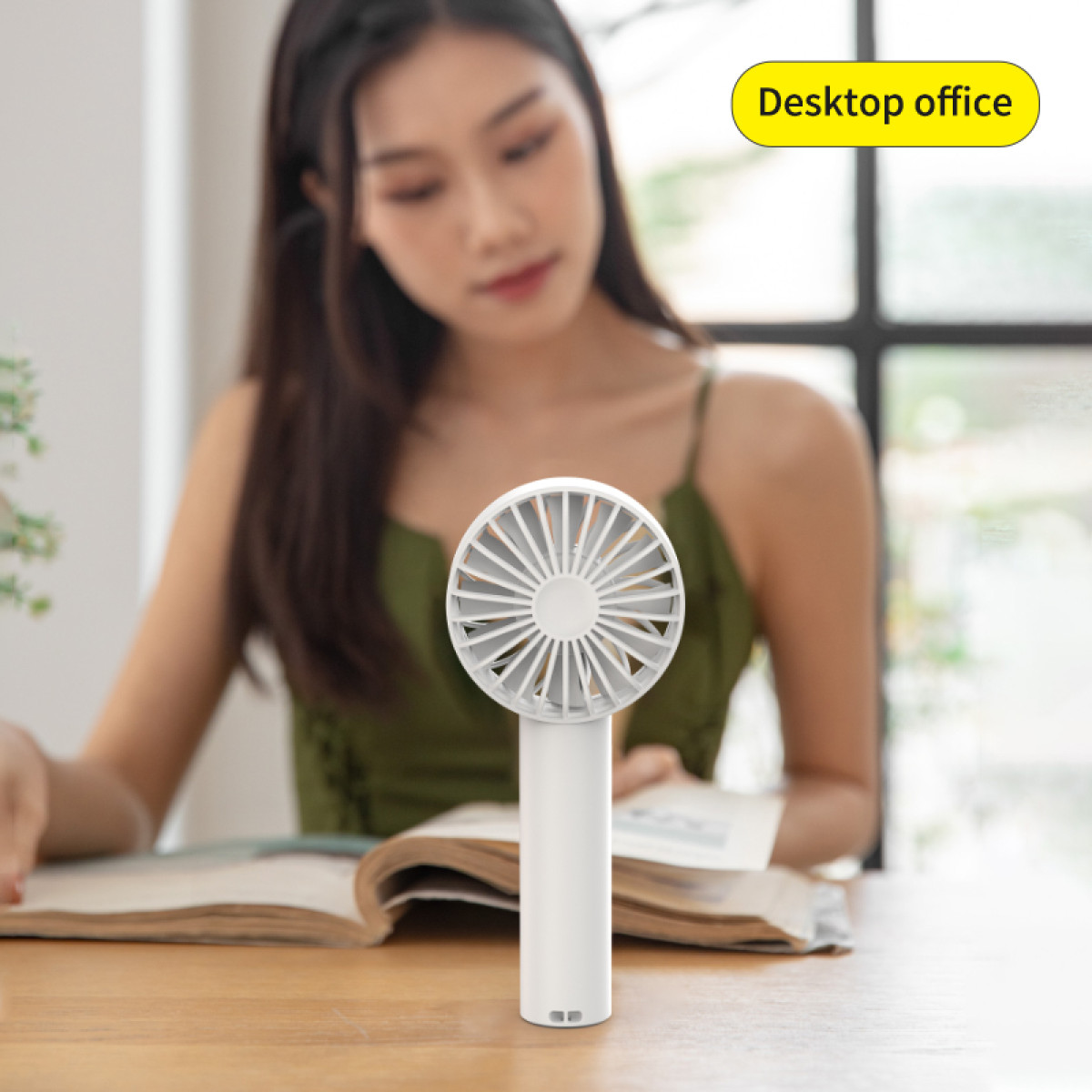 Biały wentylator ręczny na drewnianym biurku. Kobieta w zielonej bluzce czyta książkę w tle. Widoczny tekst „Desktop office”.